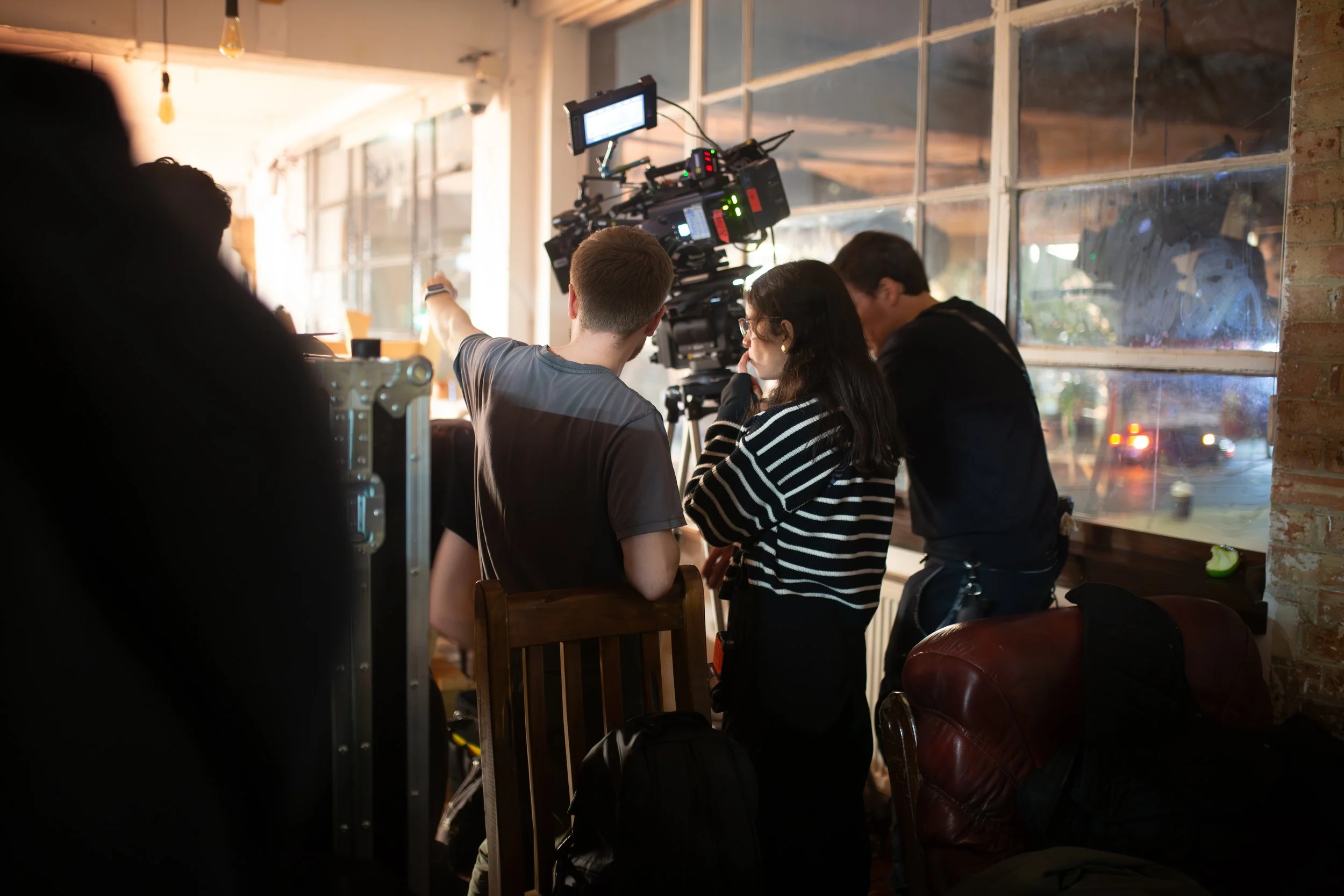 Film crew operating a camera inside a studio or cafe with large windows, brick walls, and warm lighting.