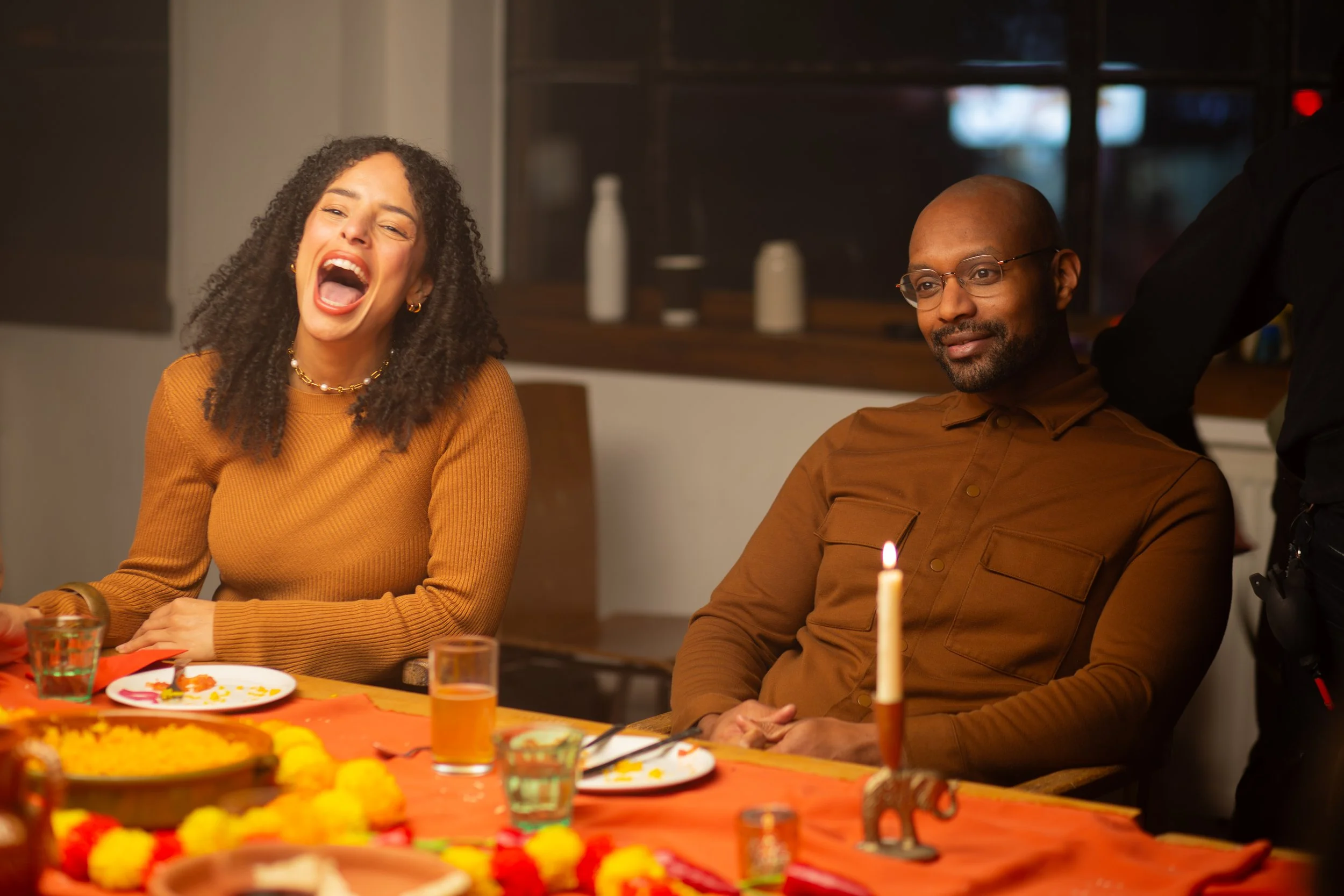 A woman with curly hair laughing and a man wearing glasses with a serious expression sitting at a decorated table with a lit candle, food, and drinks during a celebration.