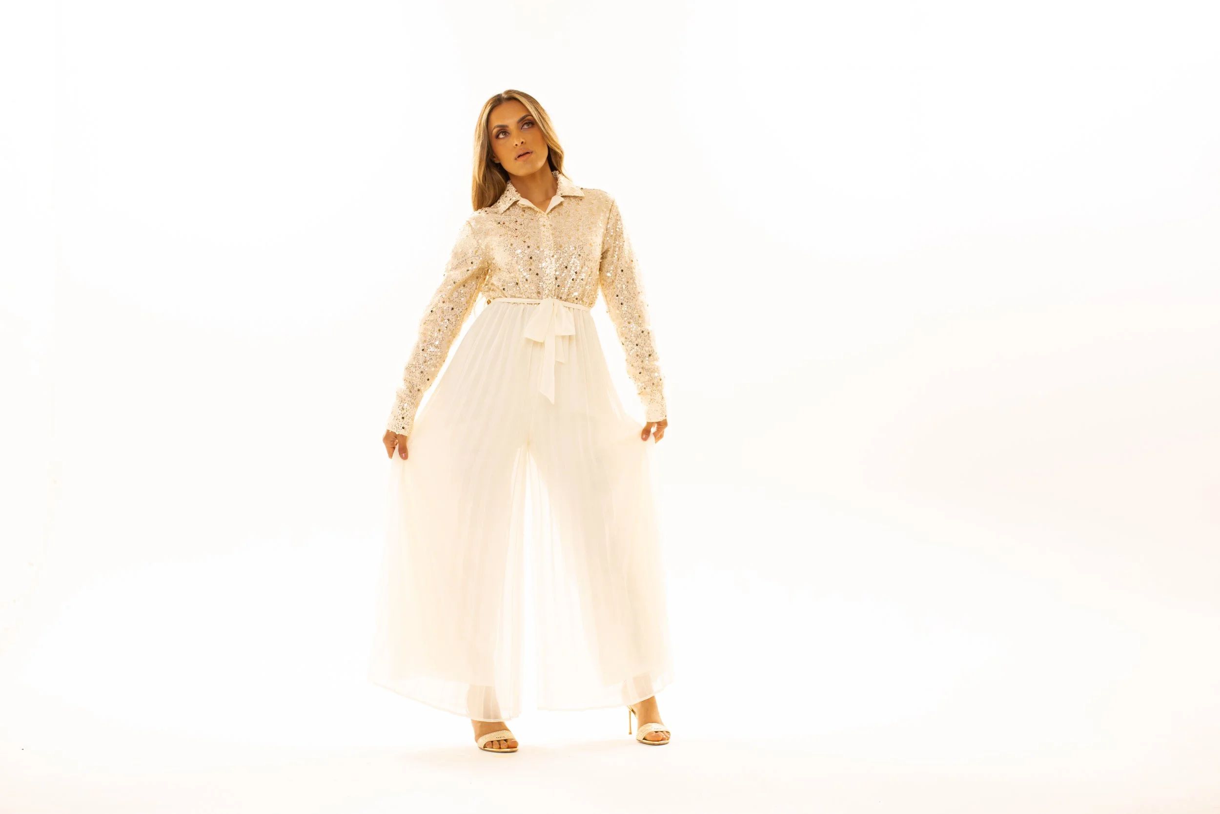 A woman with long blonde hair wearing a cream-colored sequined shirt and a flowing white skirt with a bow, standing against a white background.