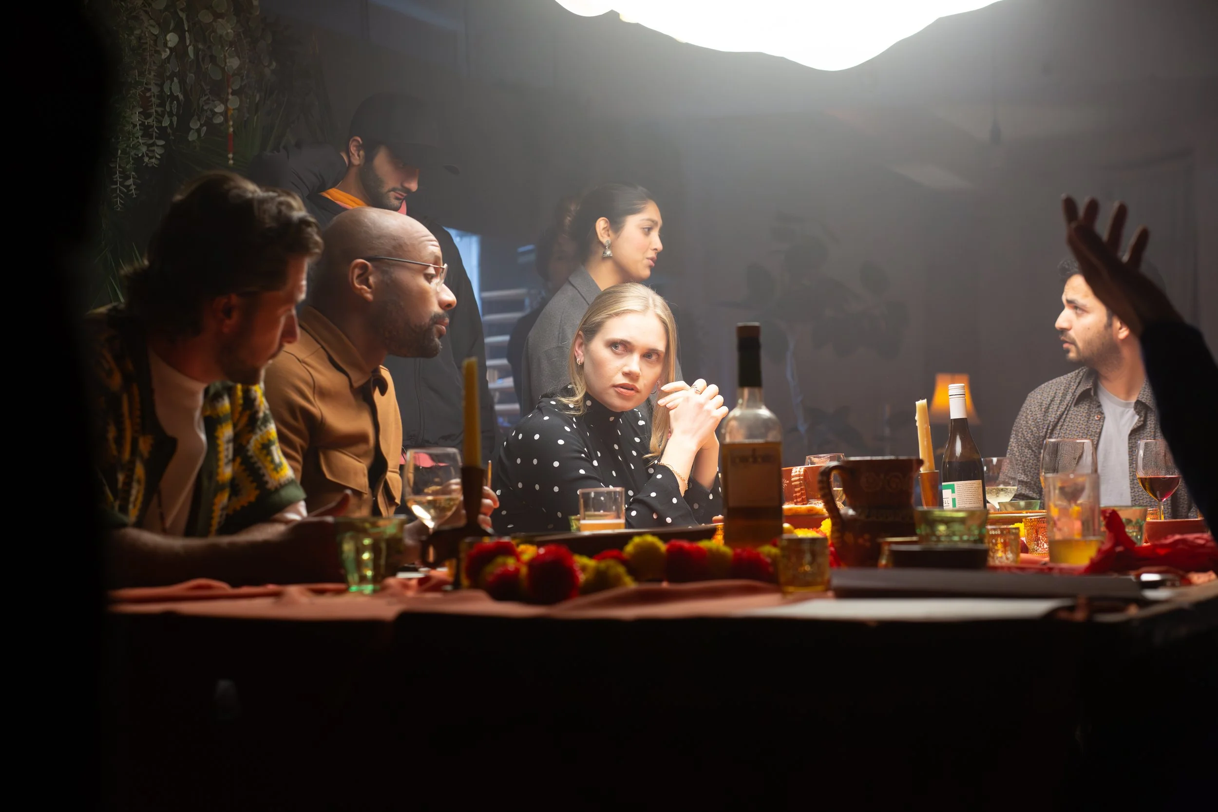 A group of people sitting and standing around a dinner table during a celebration or gathering, with candles, drinks, and flowers on the table, in a warmly lit room.