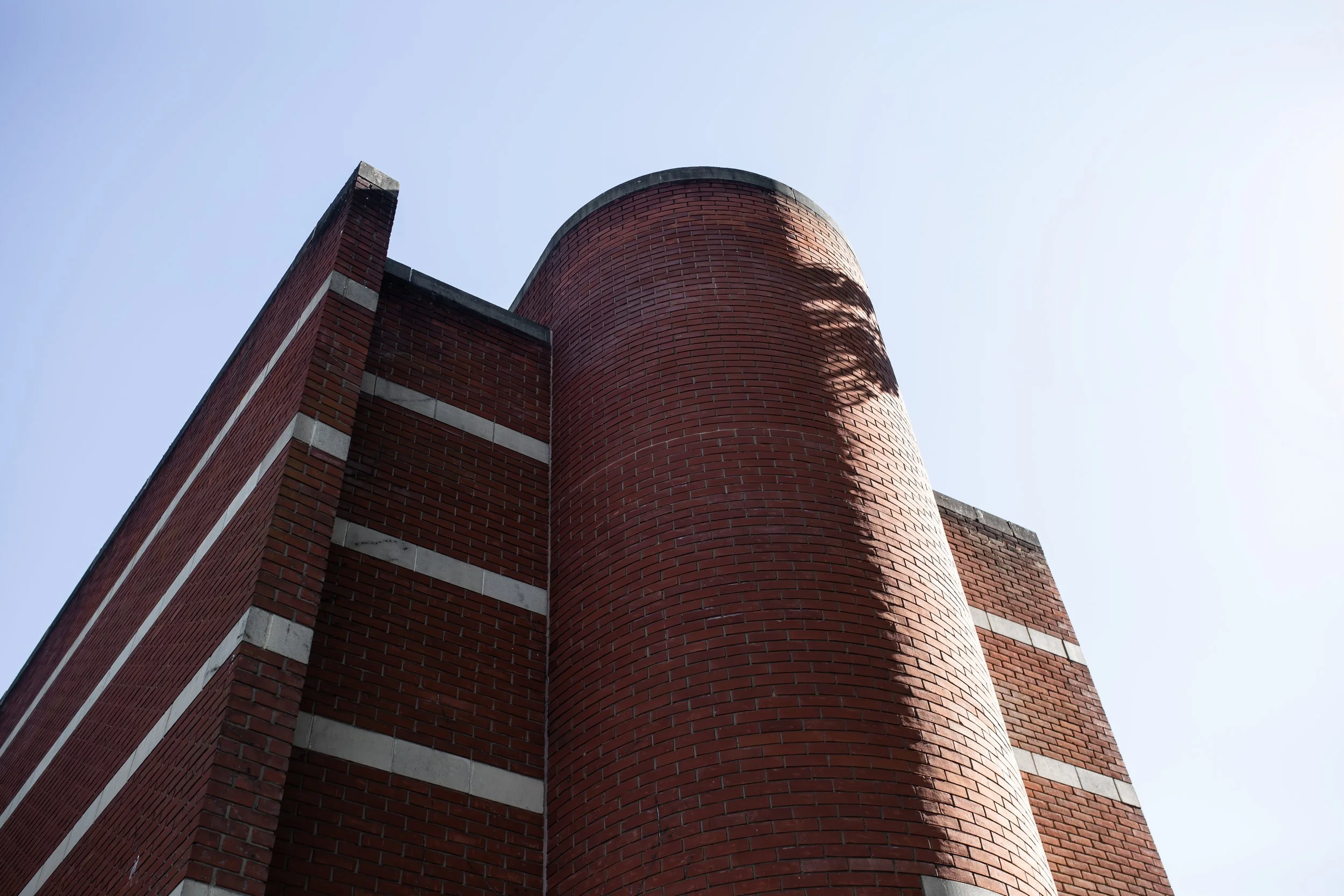 Close-up of a red brick building corner with a cylindrical chimney or structure, against a clear, light blue sky, casting diagonal shadows.