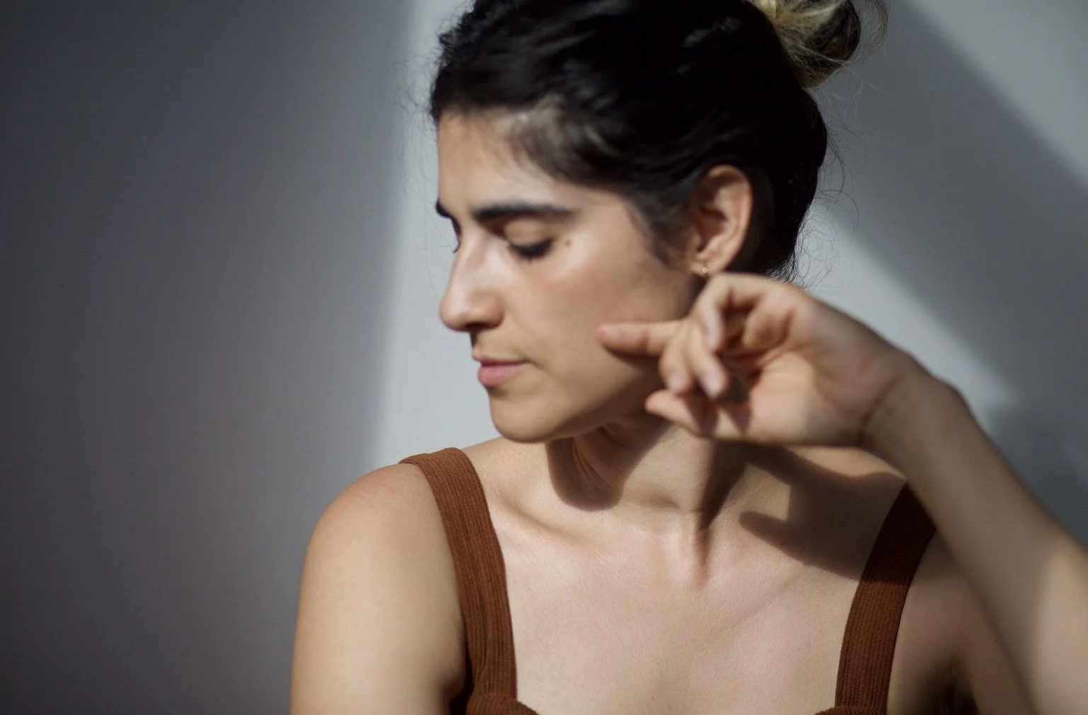 Young woman with dark hair styled in a bun, wearing a brown sleeveless top, looking down with eyes closed, touching her neck with her right hand, with neutral lighting creating soft shadows.