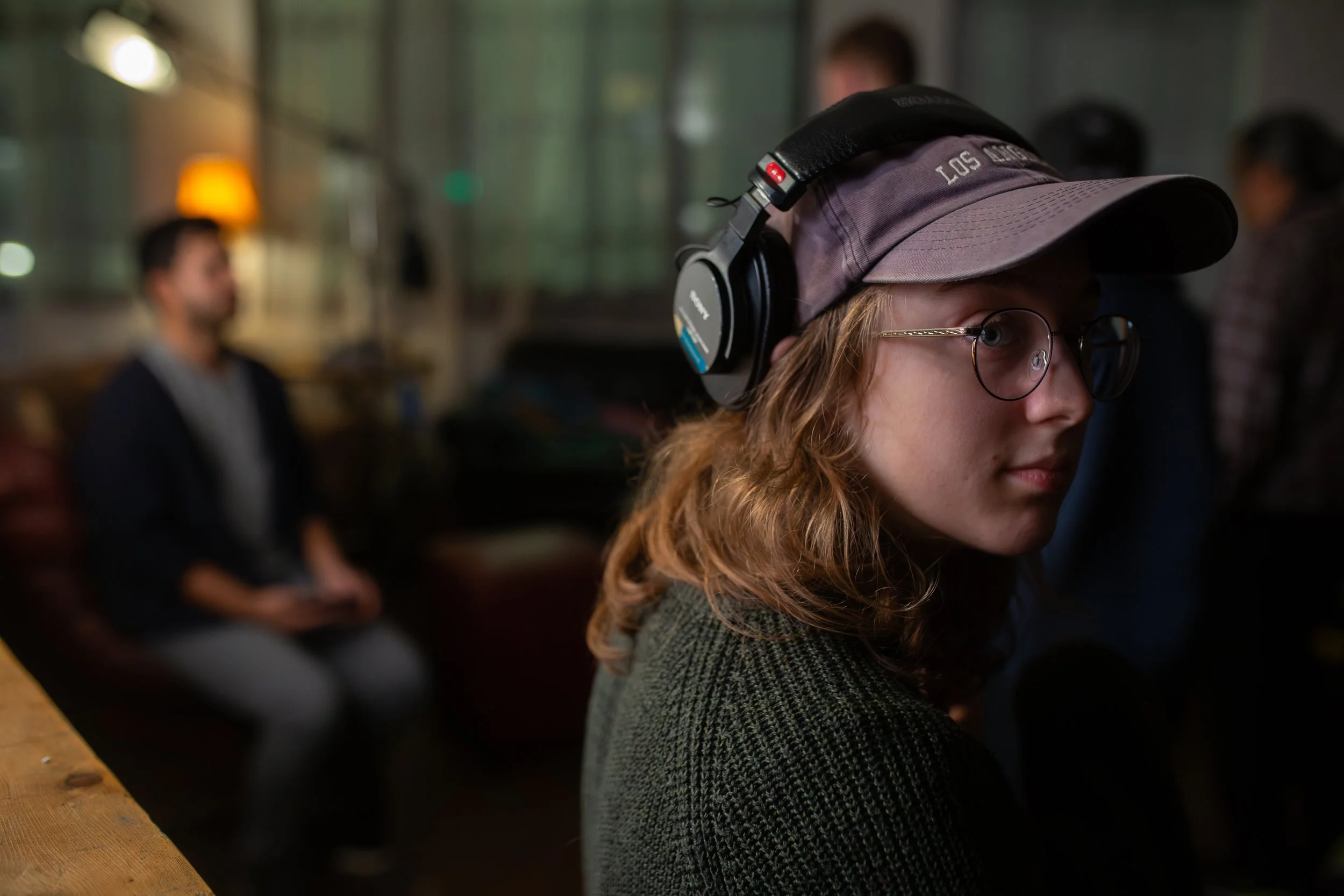 A young woman with glasses, wearing a cap and headphones, sitting indoors at a cafe or restaurant. In the background, there are other people and warm lighting.