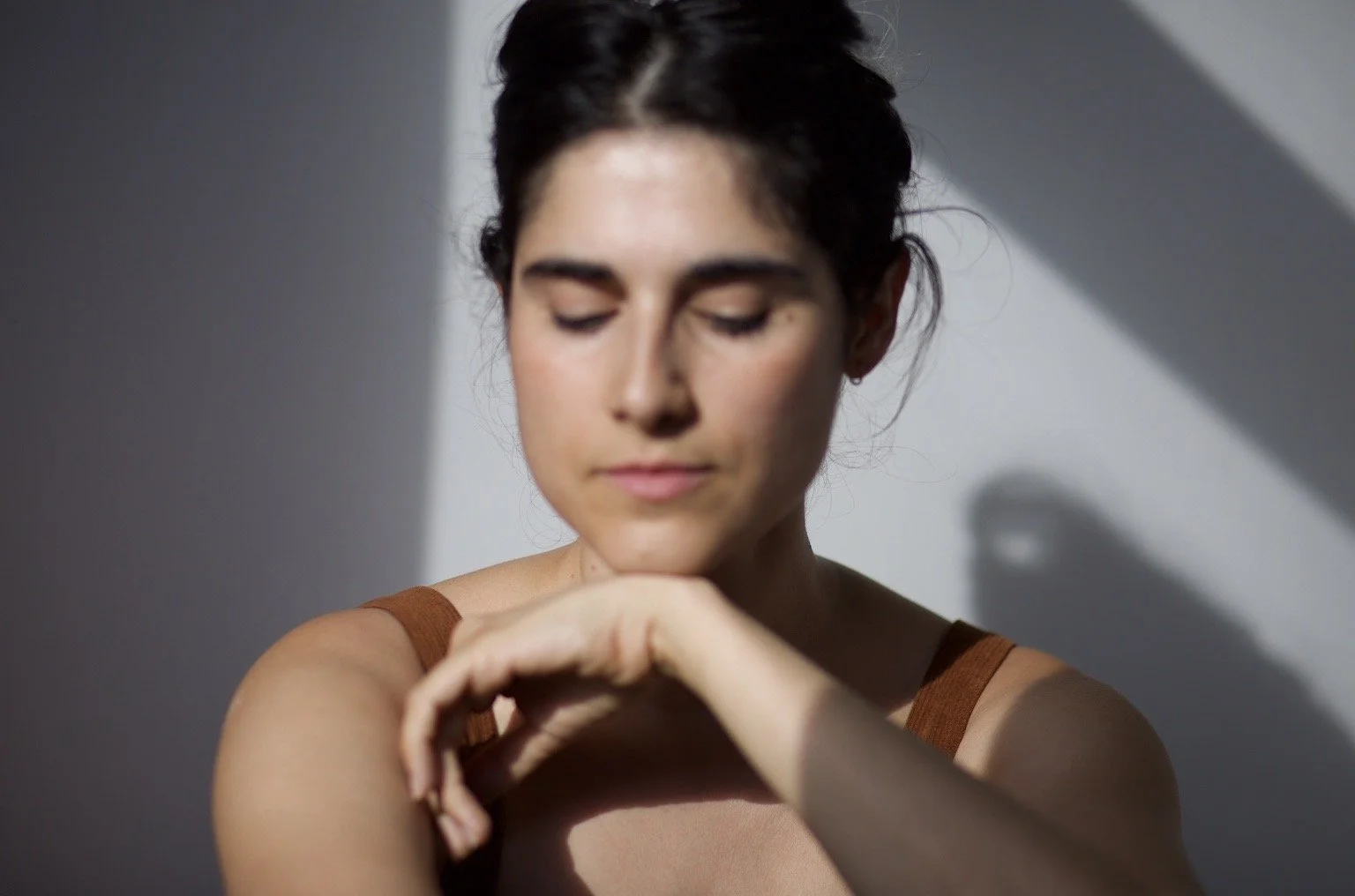 A young woman with dark hair in a bun, wearing a brown tank top, looking down with eyes closed, her right hand gently touching her left arm. Soft sunlight casting shadows on a plain light-colored wall.
