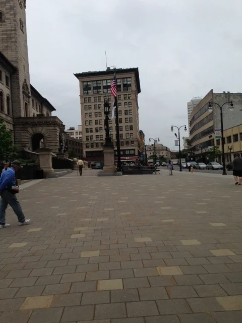 This plaza in front of city hall works well here. In fact, Dan gave this whole area an A. We have retail down here, the functions of city hall, this pedestrian plaza, the historic buildings. This is the heart... right here. Now, we just have to increase the housing so people are living here and the area does not go to sleep after the workday. The walkscore is probably high here where we are standing. His suggestion for improvement: The intersection at Main Street and Pleasant Street might be a good candidate for removing the lights altogether to provide shared space use of cars, bikers and walkers. Look up seven dials in England, he suggested.