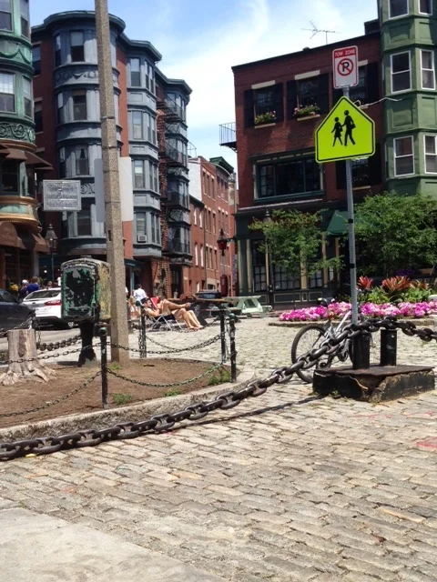 This photo was taken directly across from the Paul Revere House. This is a neighborhood for walkers and bikers as you can clearly see from the "no parking" sign above the image of walkers and the bicycle tied to the pole below. Check out the sunbathers in the pedestrian plaza in the center of the square and the streets lined with old brick cobblestones. All throughout this dense, mixed use neighborhood, you will see the storefronts on the first floor and the residential units above.