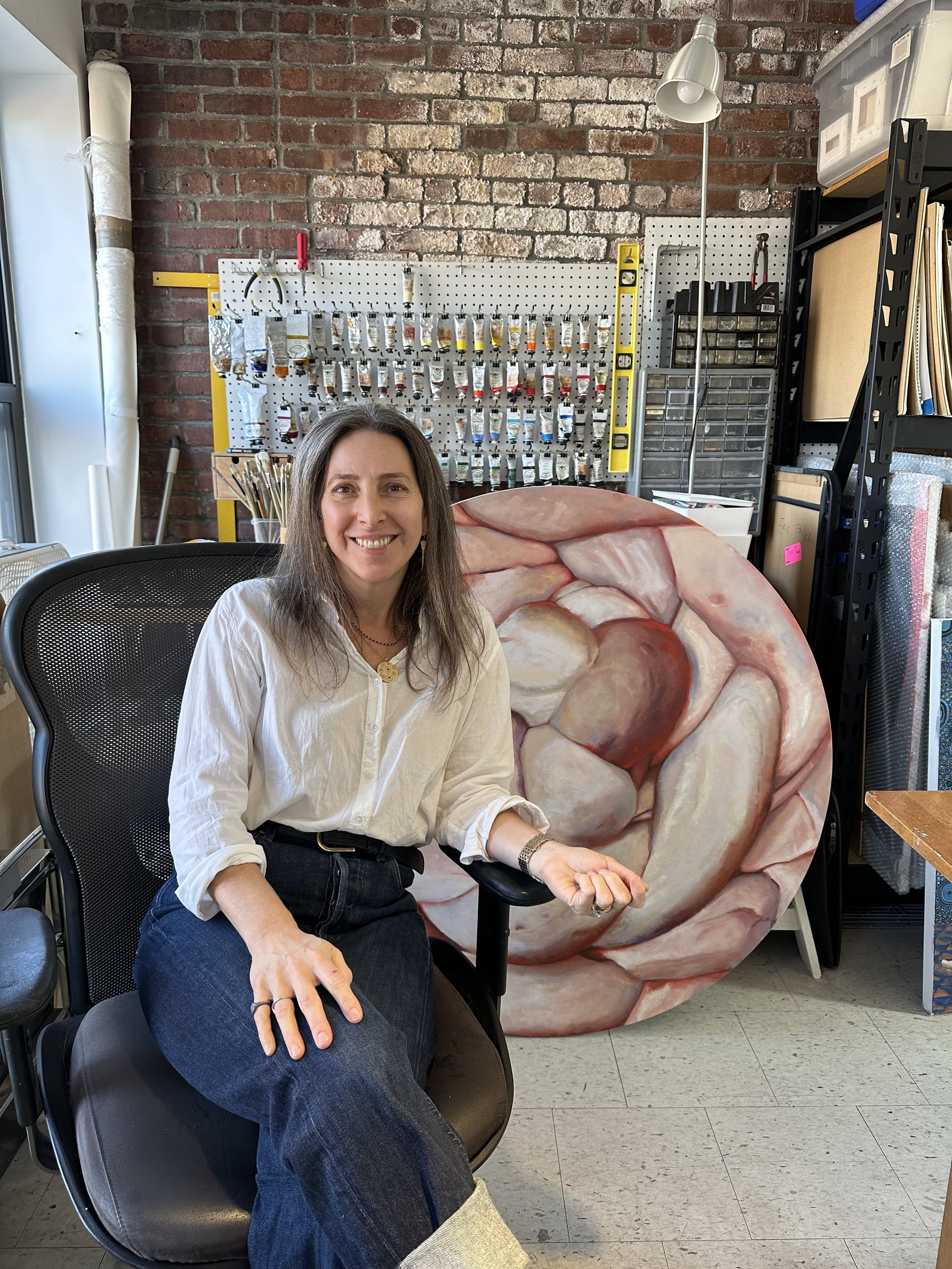 A woman sitting in a black office chair, smiling, in an art studio. Behind her, there is a large circular painting of a flower, a pegboard with art supplies, and storage shelves with various materials.