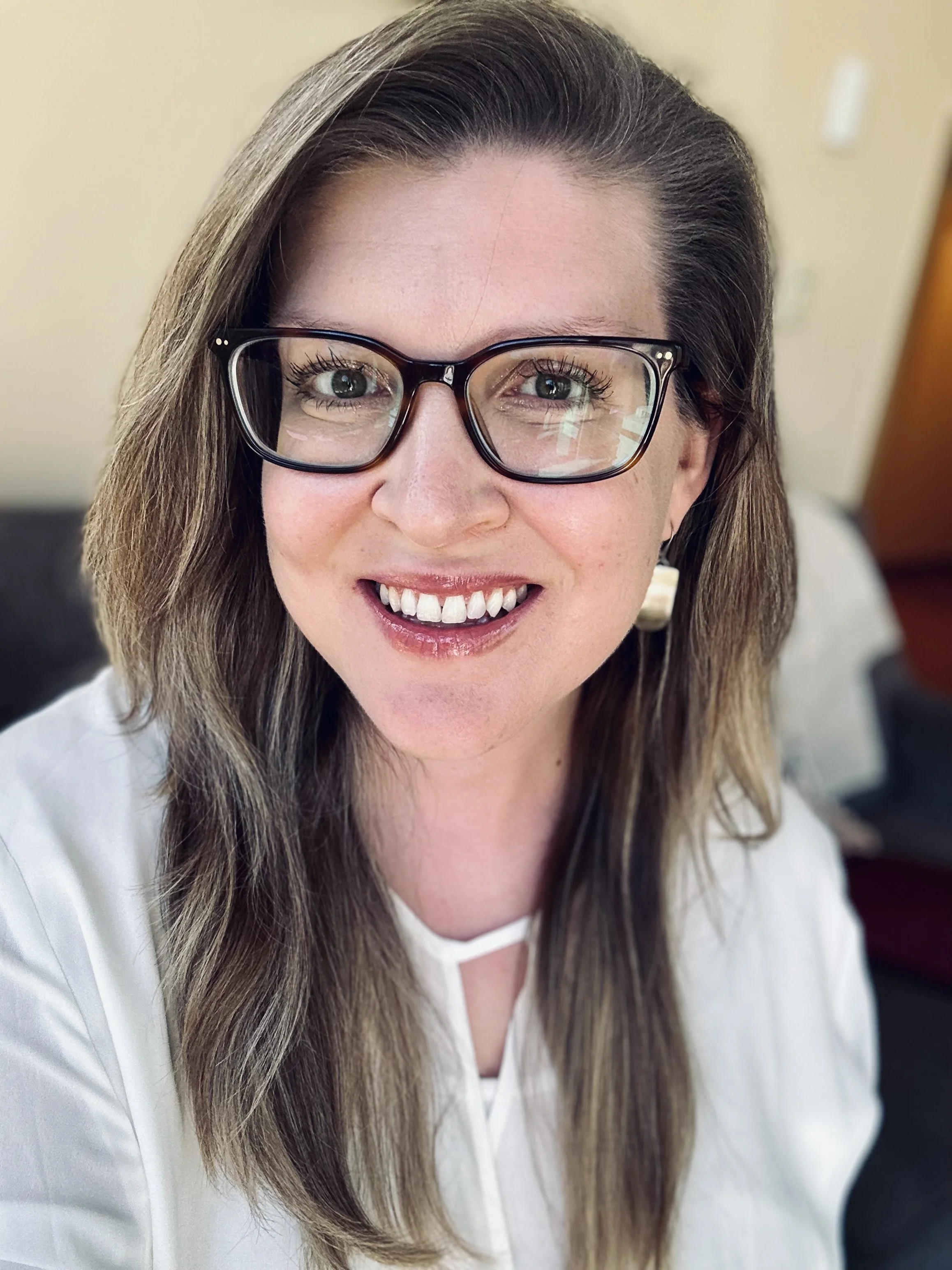 A woman with long brown hair, wearing black-framed glasses, smiling, dressed in a white top, sitting indoors.