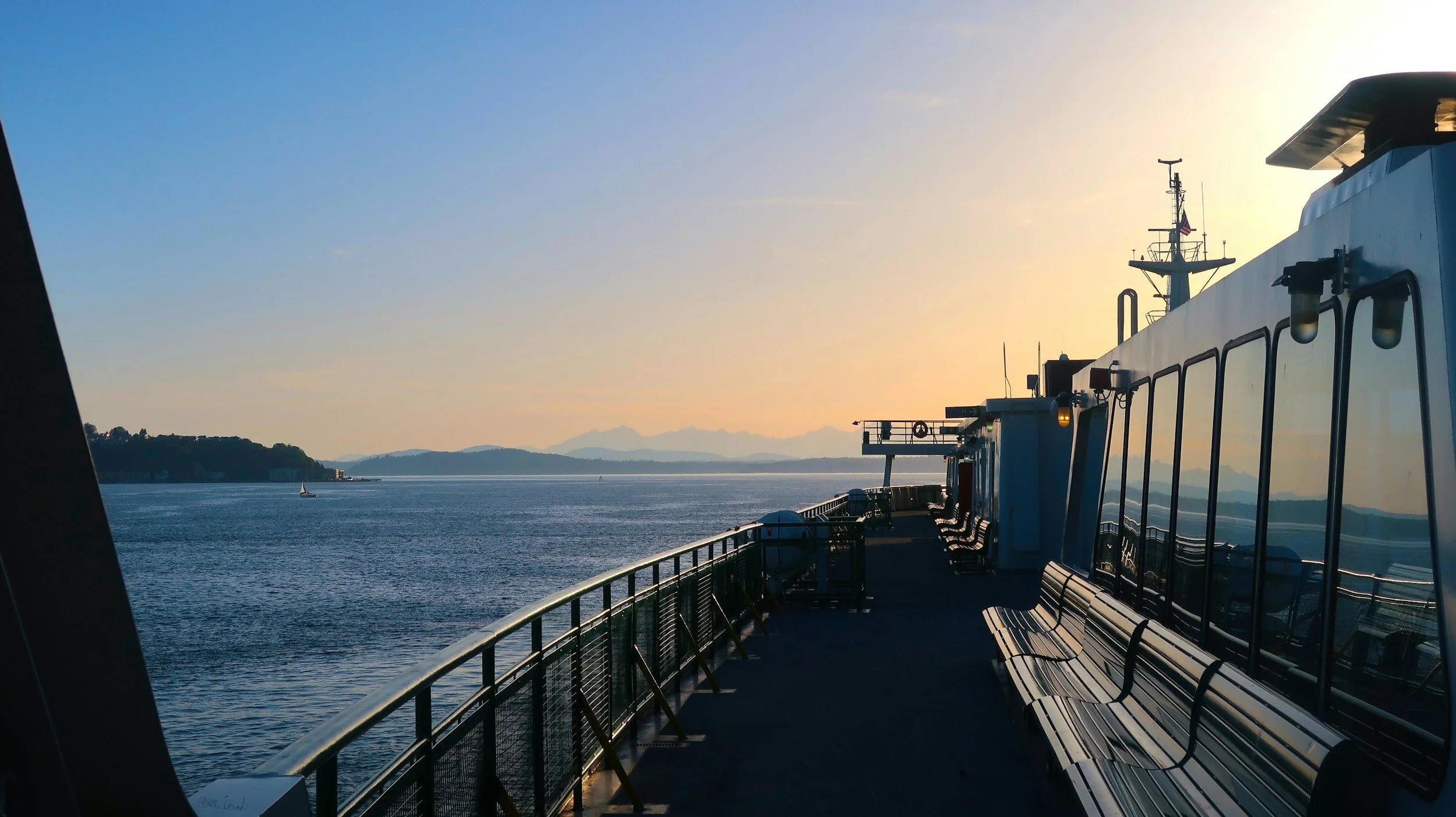 View from the deck of a ferry or ship, with empty benches and a railing, looking out onto calm water and distant mountains at sunset.
