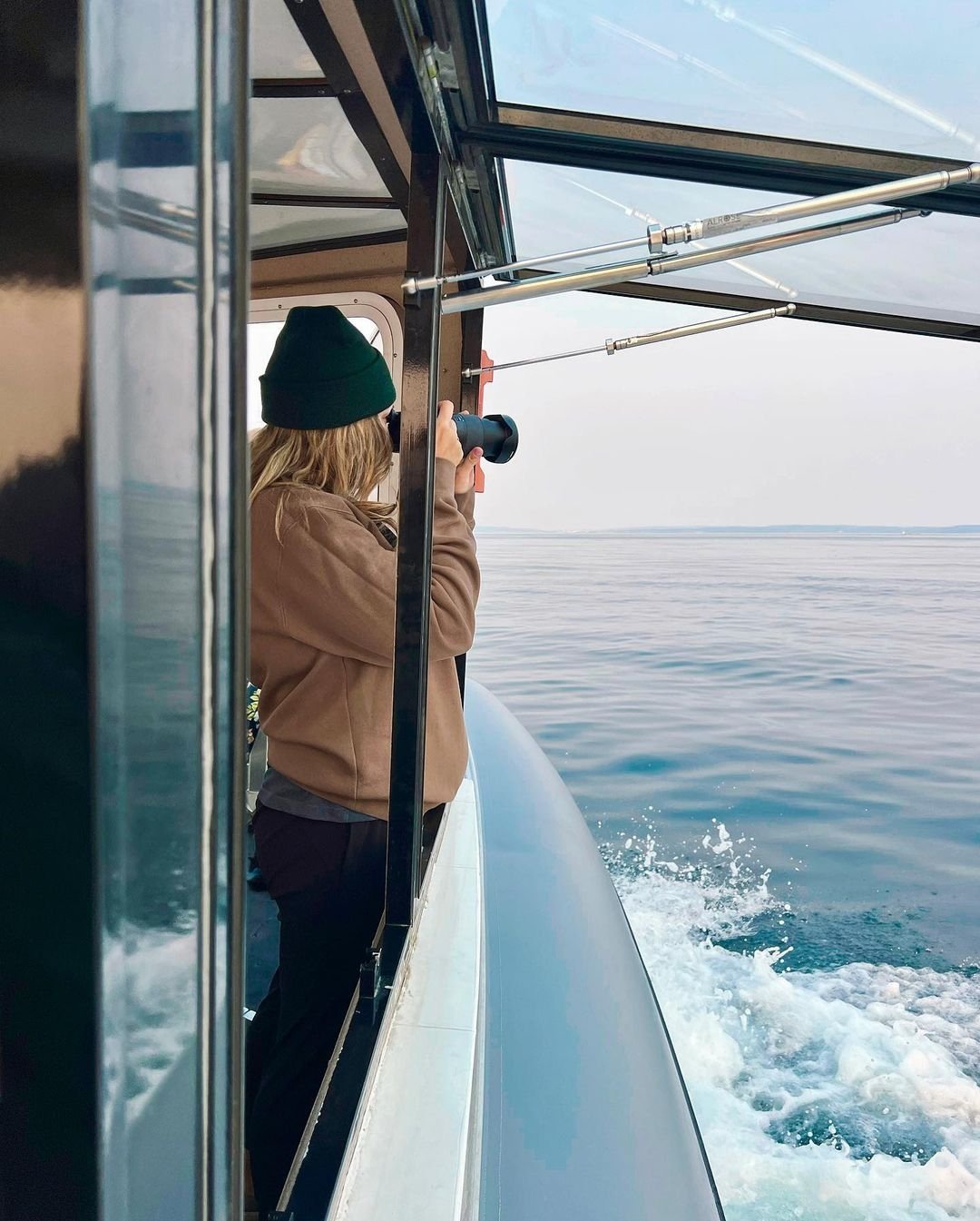 Person with blonde hair, wearing a green beanie and brown jacket, taking photos with a camera on a boat.