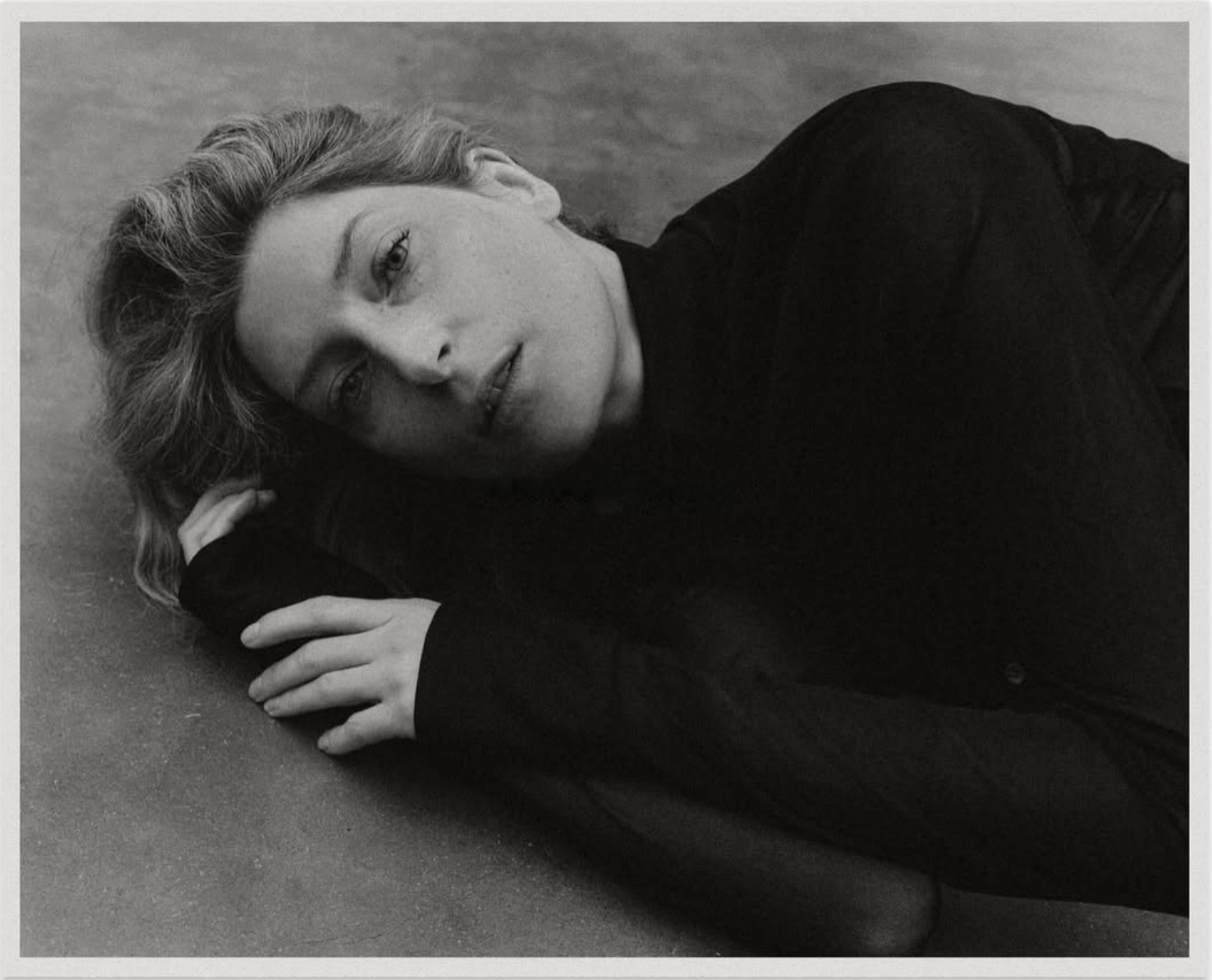A woman lying on the floor, resting her head on her hand, looking at the camera. She has short, wavy hair and is wearing a dark top.