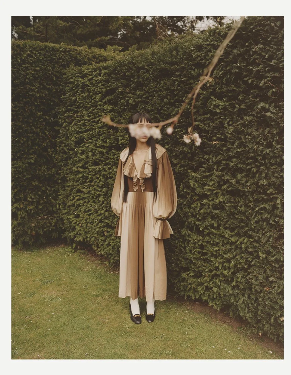 A woman in a vintage-inspired tan and beige outfit standing outdoors in front of a dense green hedge, with a branch with pink blossoms hanging in front of her face.