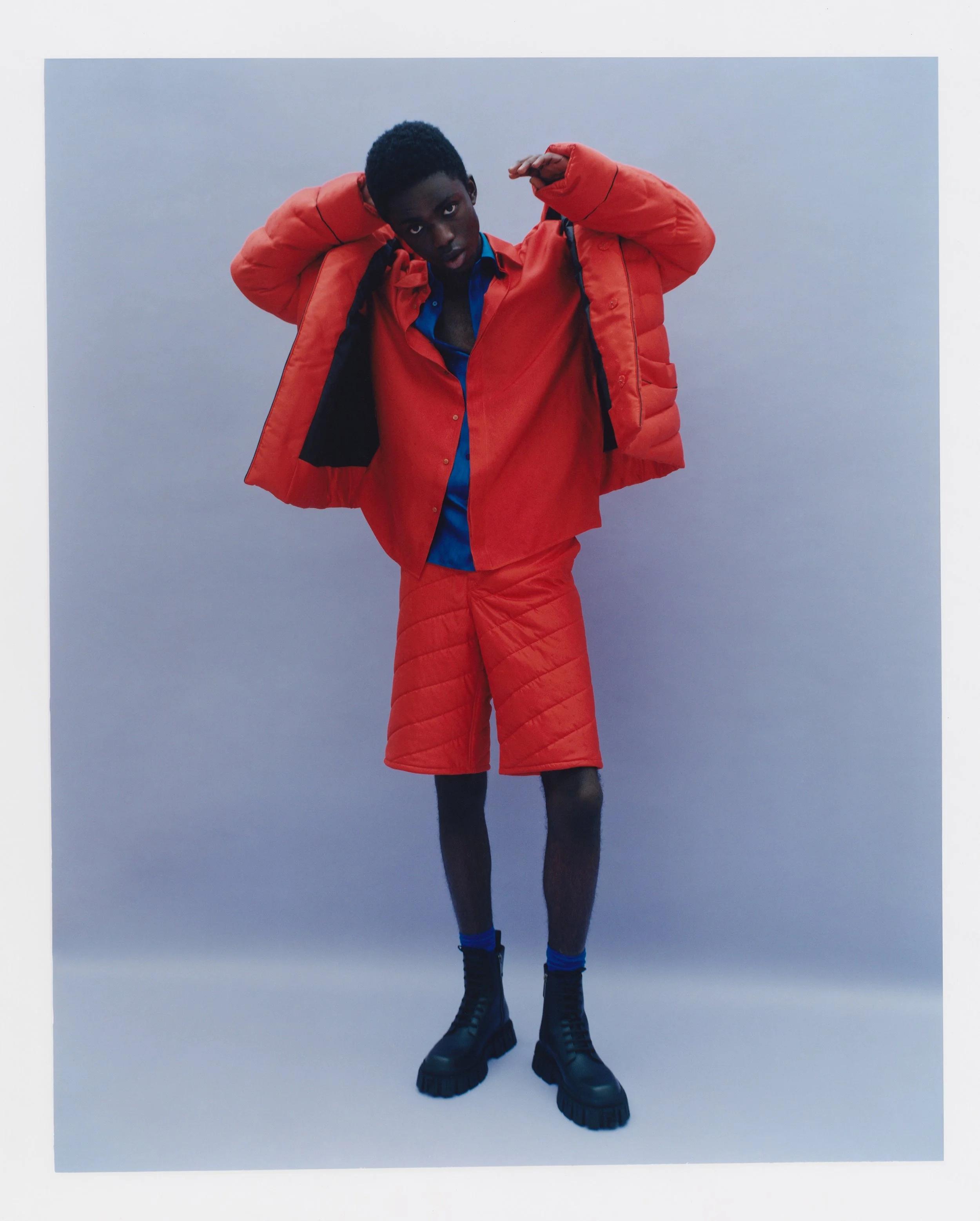 A young man with dark skin and short black hair adjusting a bright orange puffy jacket over a blue shirt. He stands against a plain, light-colored background, looking directly at the camera with a serious expression. His hands are raised to his head, and he is wearing black boots and blue socks.