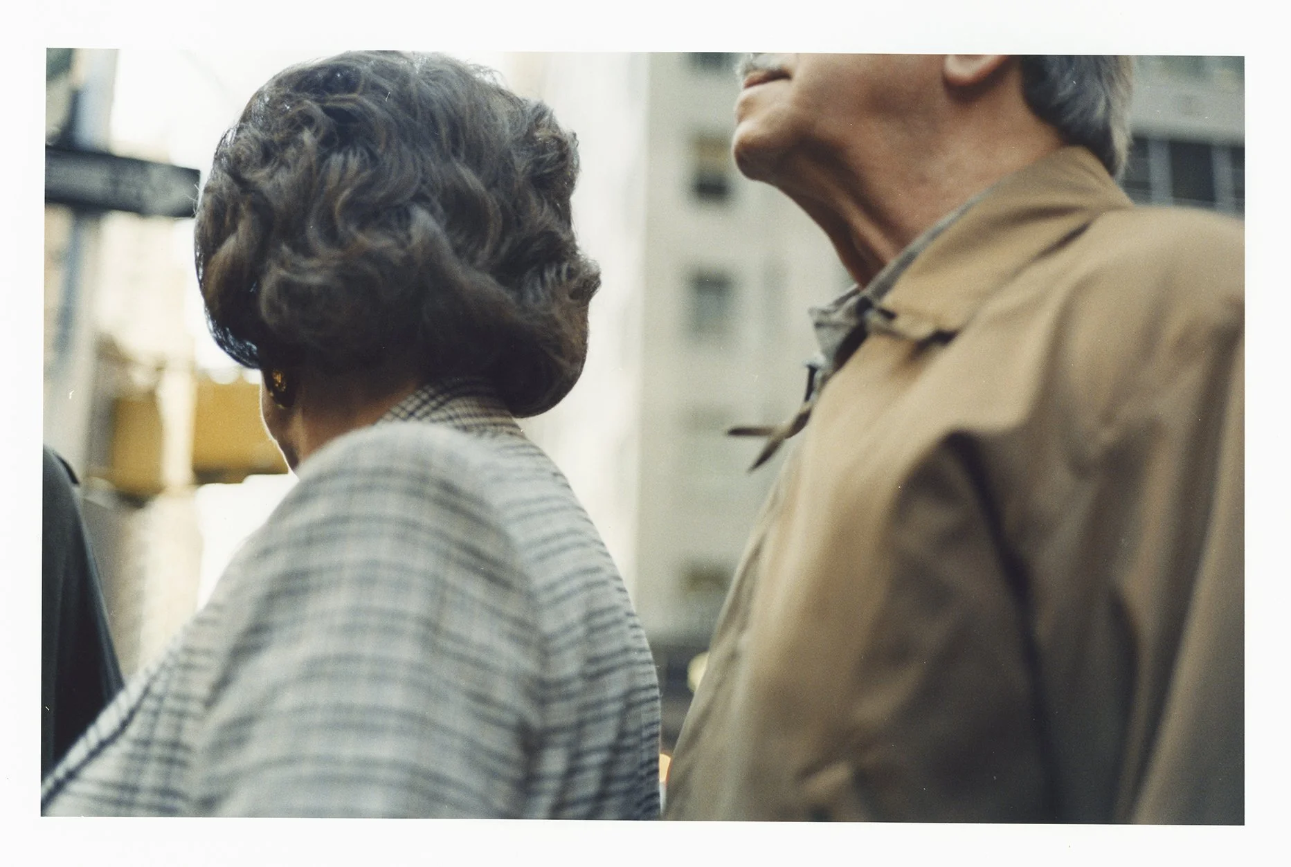 Close-up of two elderly individuals, a woman and a man, standing close together on a city street, with the woman wearing a checked blazer and earrings, and the man dressed in a brown jacket.
