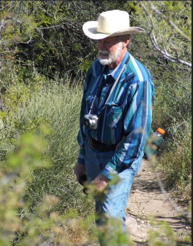 Ranch owner stands in overgrowth with hat and camera.