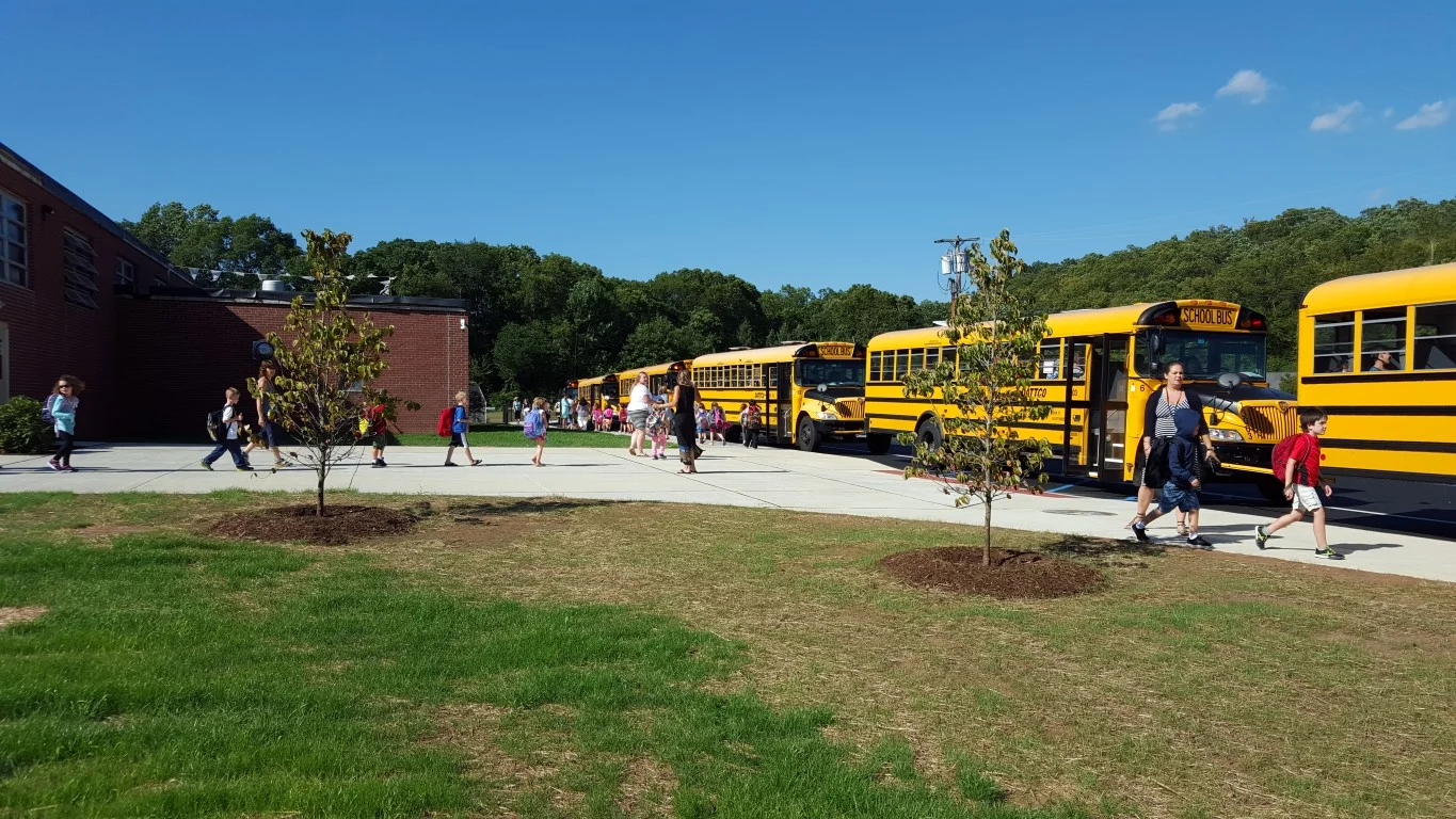 Eleven buses lined up at once in the dedicated bus lane while kids walk on the sidewalk and no longer crossing the driveway.