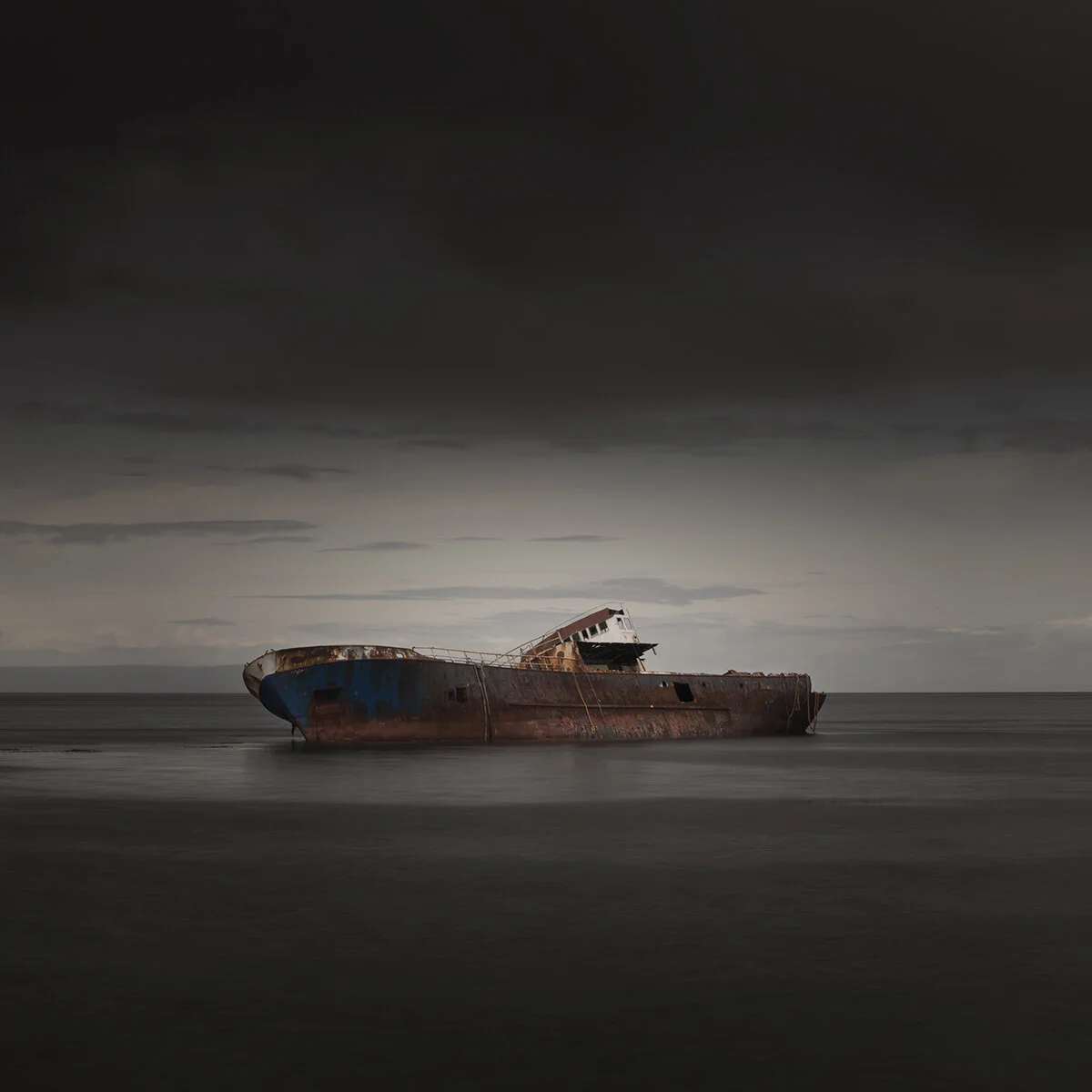 A rusted, abandoned shipwreck in calm water under a cloudy, dark sky.