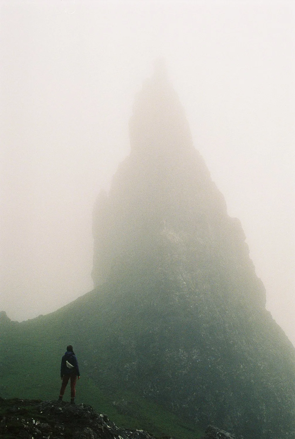 A person standing on a dark rocky surface, looking at a large, fog-covered mountain or rock formation in the background.