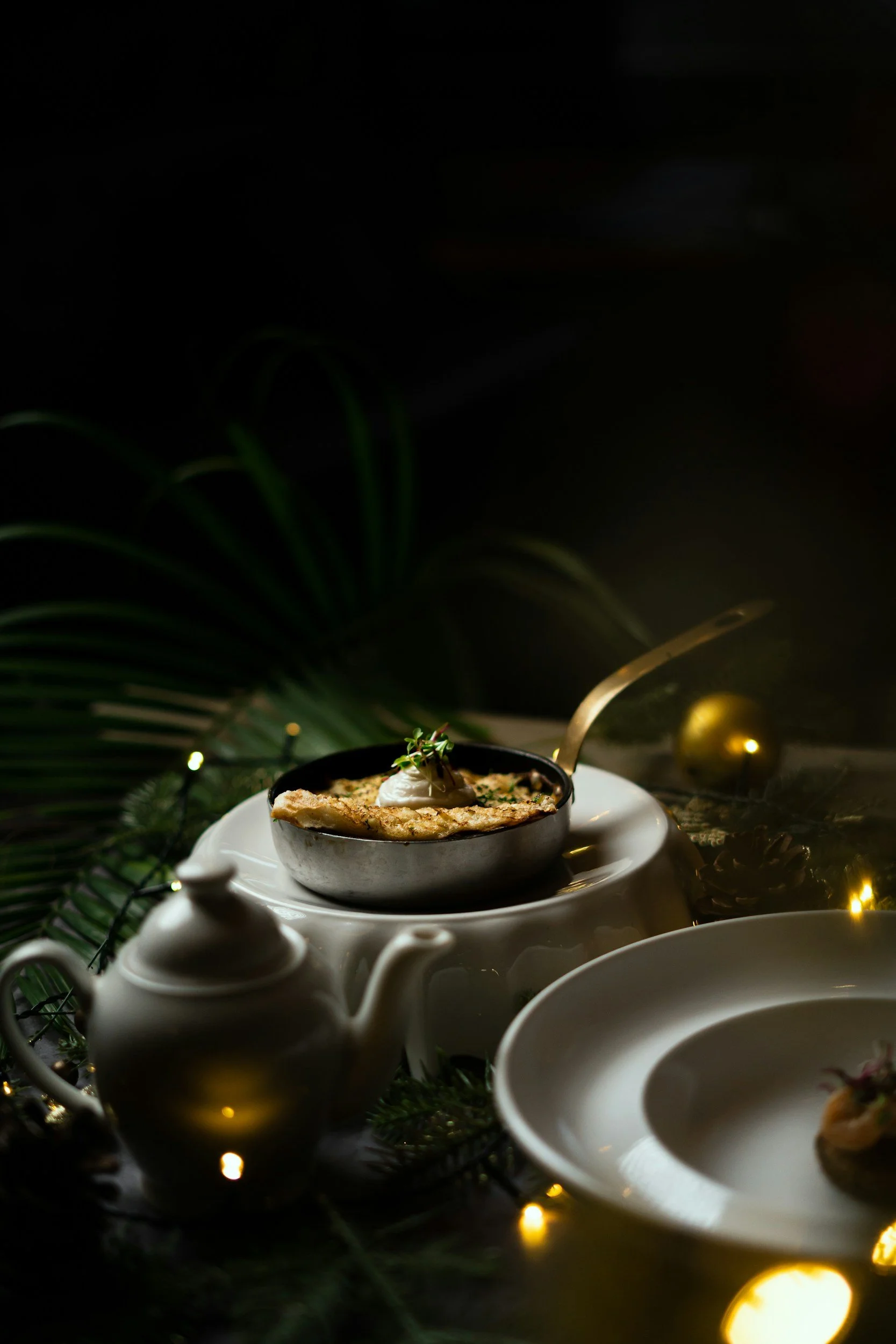 A plate of food on a table with a teapot and tea cup, surrounded by decorative greenery and warm yellow lights, with dark background.