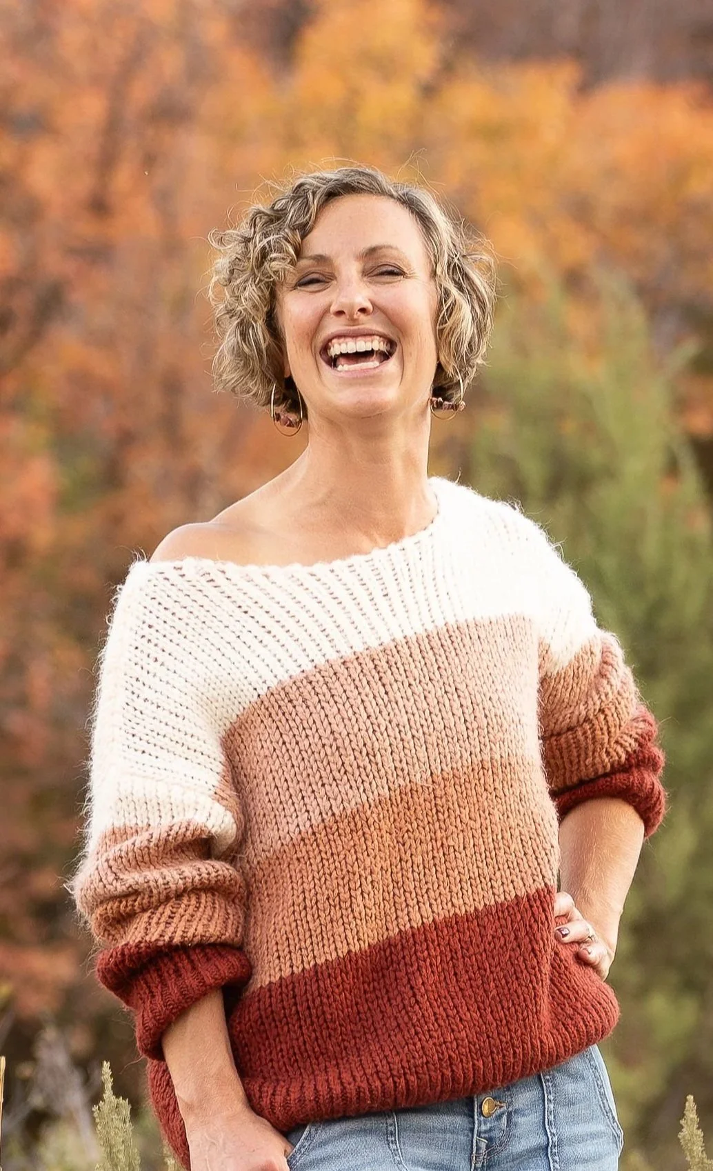 Liesel has curly short hair and is wearing a color-blocked knitted sweater while standing outdoors in front of autumn trees, smiling.