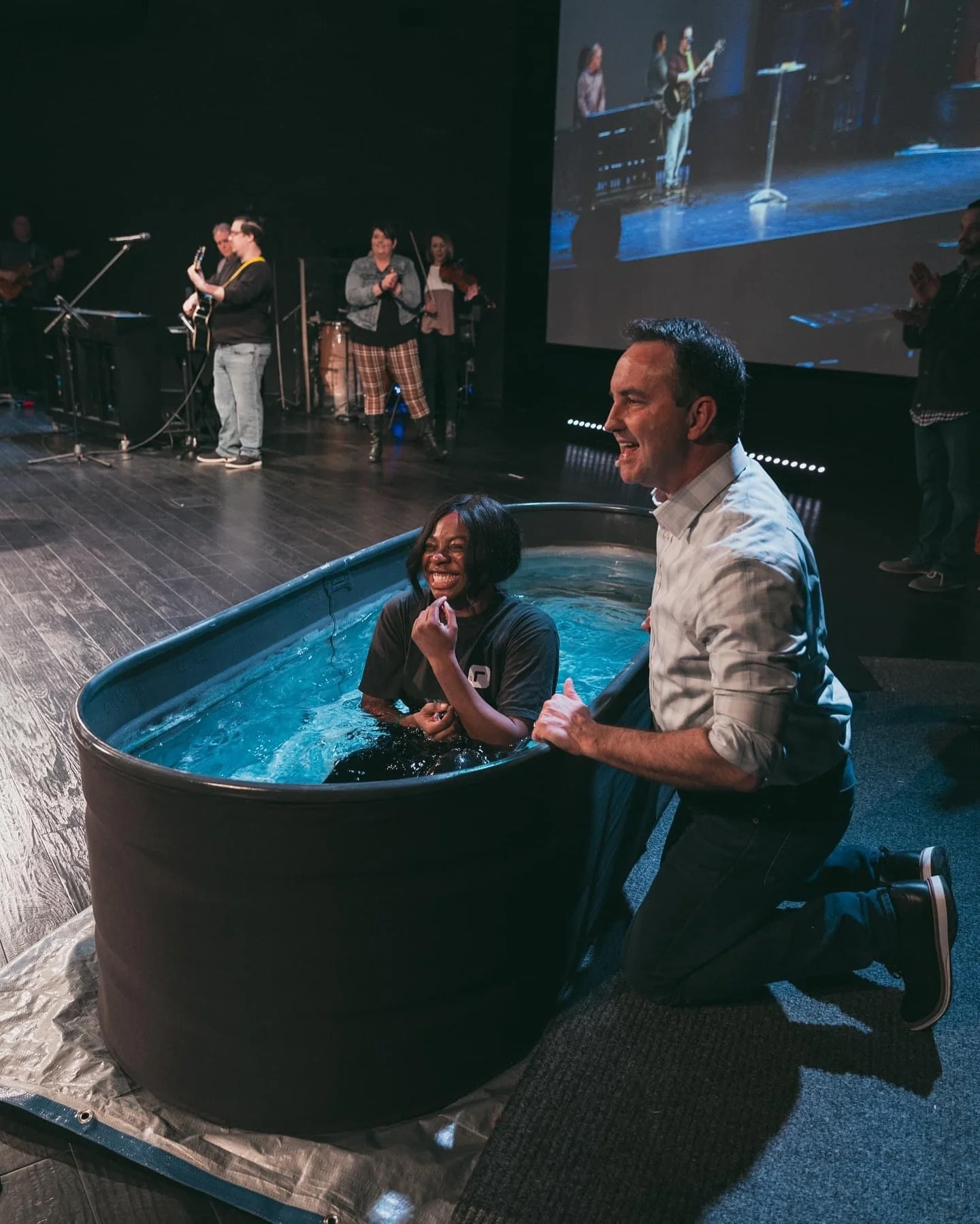A woman is sitting in a bathtub filled with water on stage, smiling and laughing. A man kneels beside her, holding onto the tub and smiling. In the background, there is a band performing on stage with several musicians playing instruments. A large sc