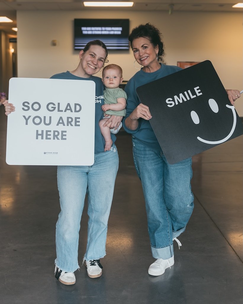 Two women and a baby standing inside a building, smiling at the camera. The woman on the left holds a sign that says 'So glad you are here' and the woman on the right holds a black sign with a large smiley face and the word 'Smile'.
