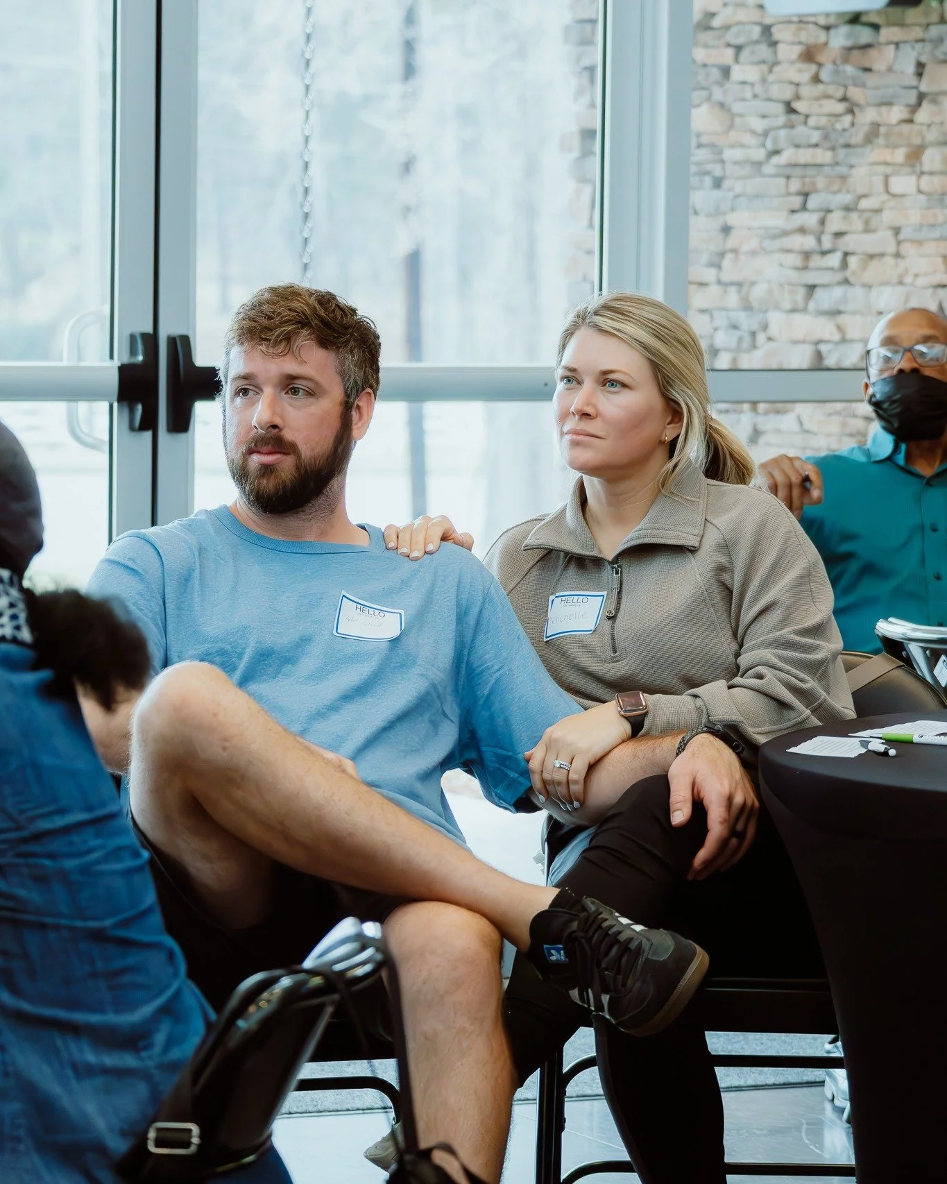 Group of people sitting indoors, participating in a discussion or workshop. One woman has her hand on a man's shoulder, and another man is in the background wearing a face mask.