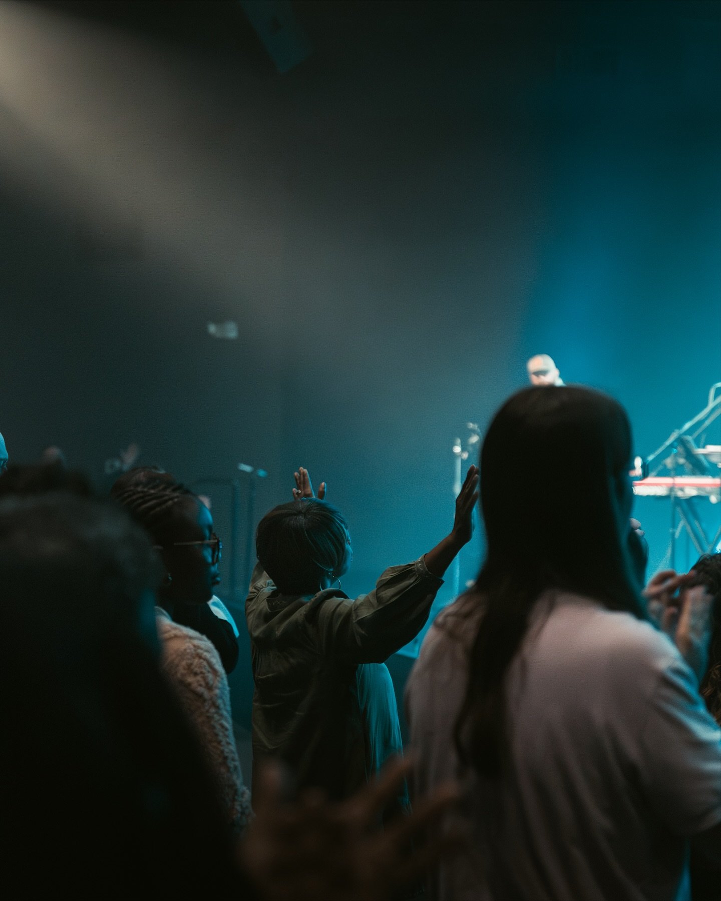 Audience at a concert with some people raising their hands towards the stage, where a performer wearing sunglasses is visible under blue stage lighting.