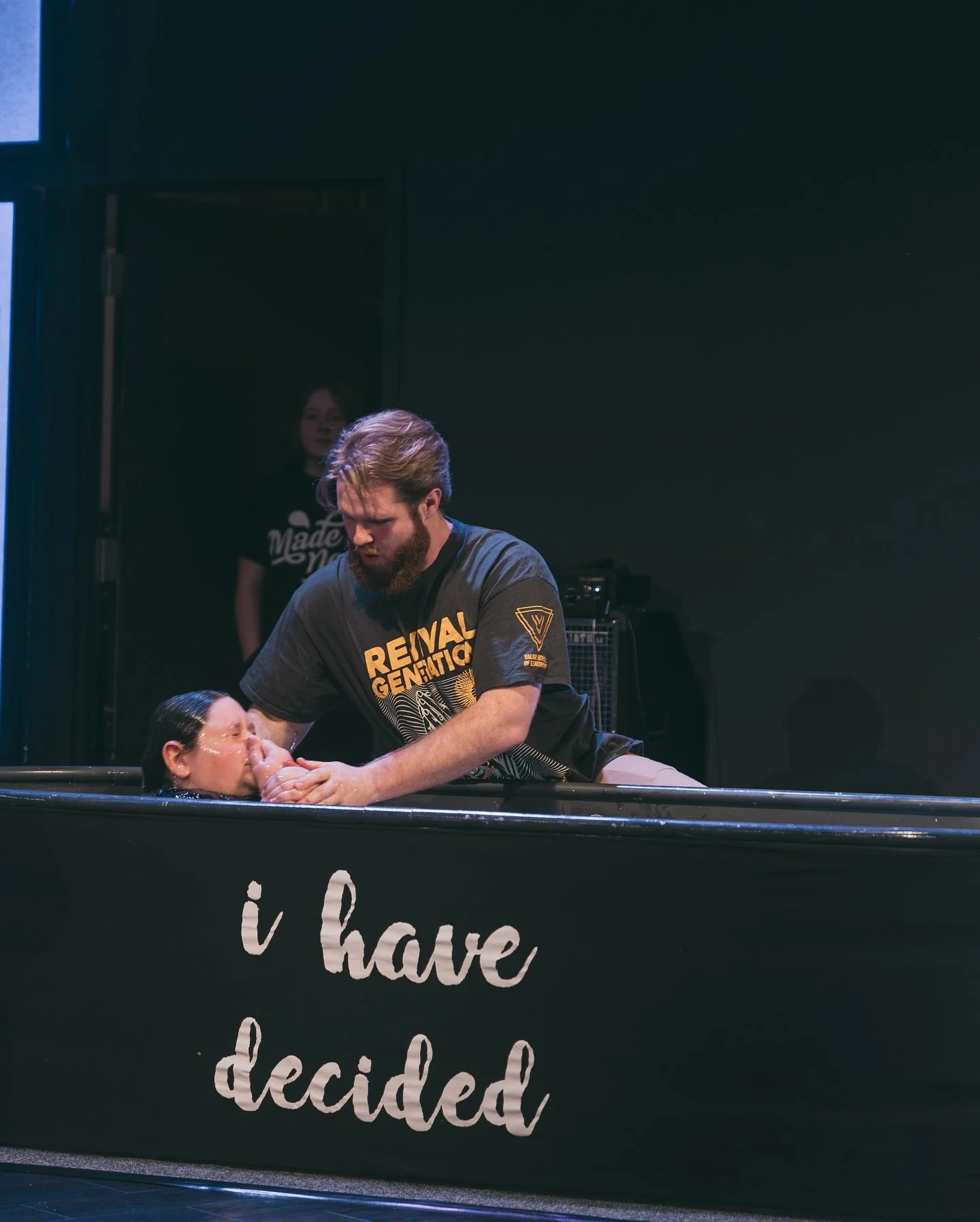 A man baptizing a woman in a baptismal pool with the words 'i have decided' written on the front, while a woman in the background watches.