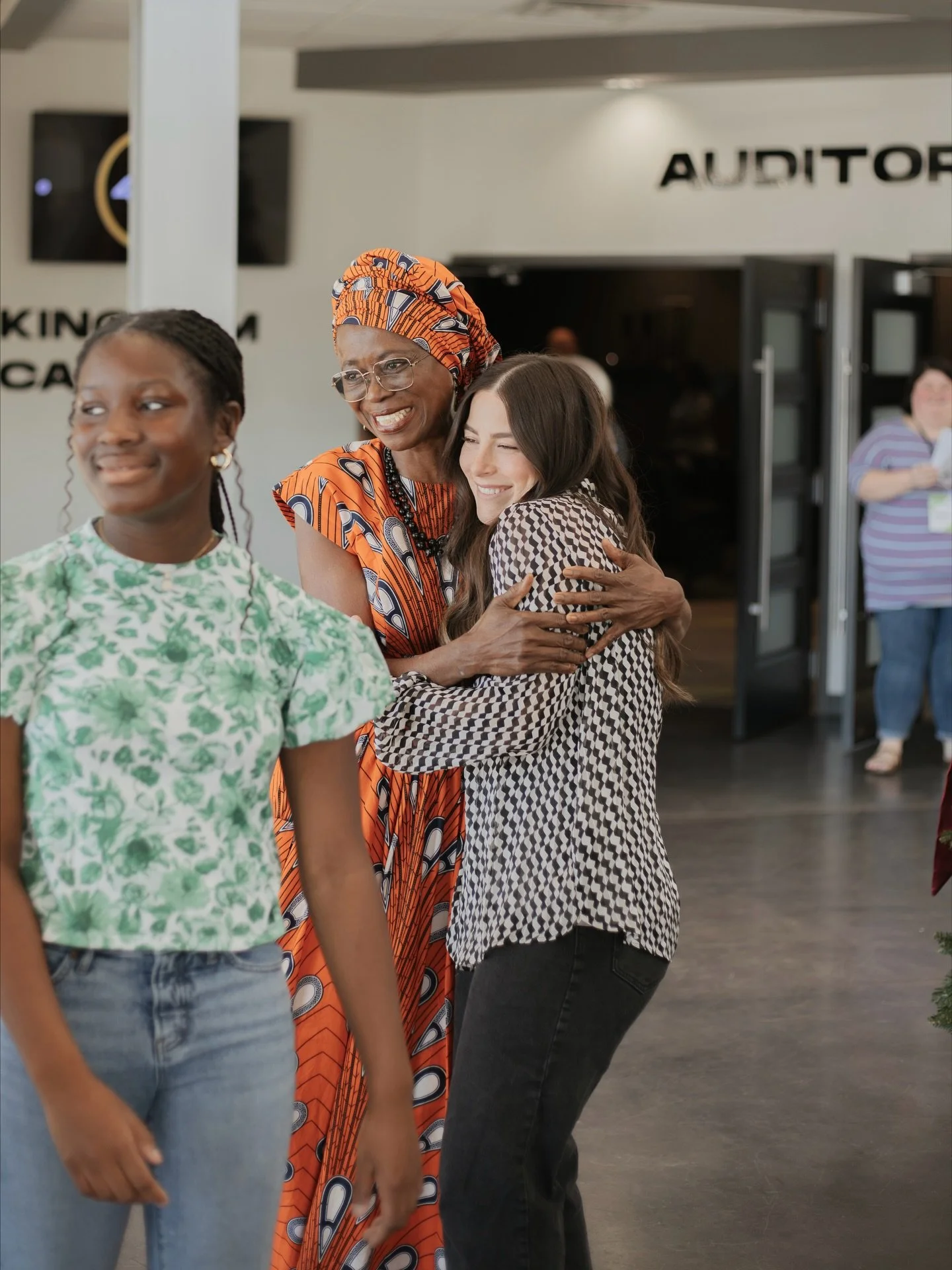 Three women embrace and smile in a public indoor space. The woman in the center wears glasses, a colorful patterned dress, and a matching headwrap. The woman on the right has long dark hair, a black and white patterned shirt, and black pants. The woman on the left has braided hair, earrings, a green and white floral top, and jeans.