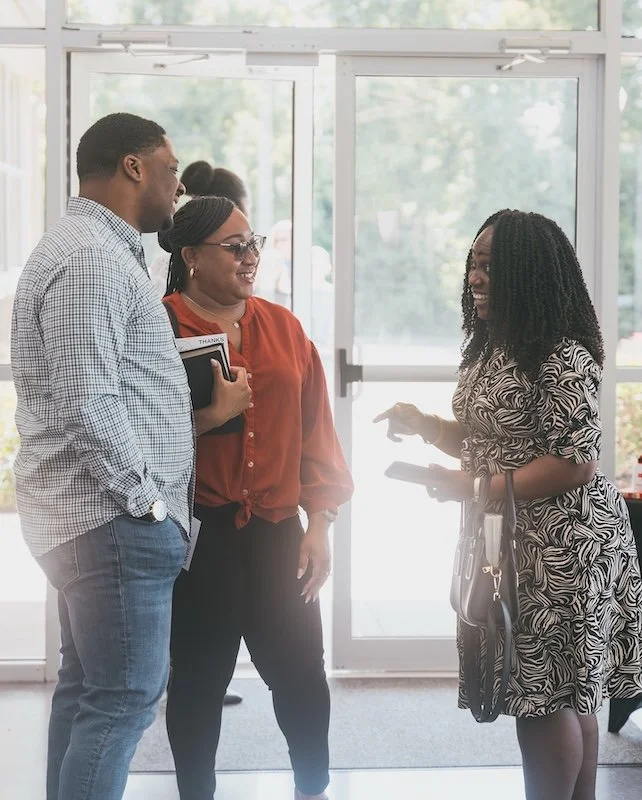 Three people, two women and one man, are standing and talking inside a building near the entrance. The woman on the right is holding a phone and a purse, while the other two are smiling and holding a book.