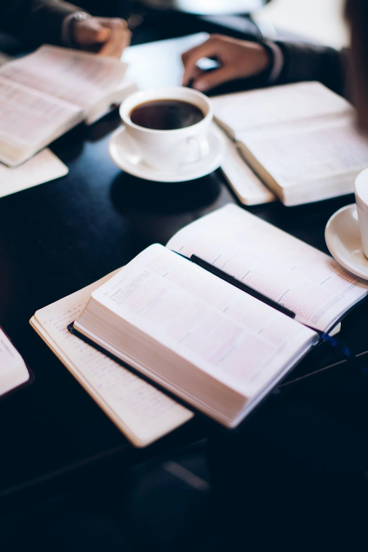 Open Bible on a table with notebooks and cups of coffee in a study or meeting setting.
