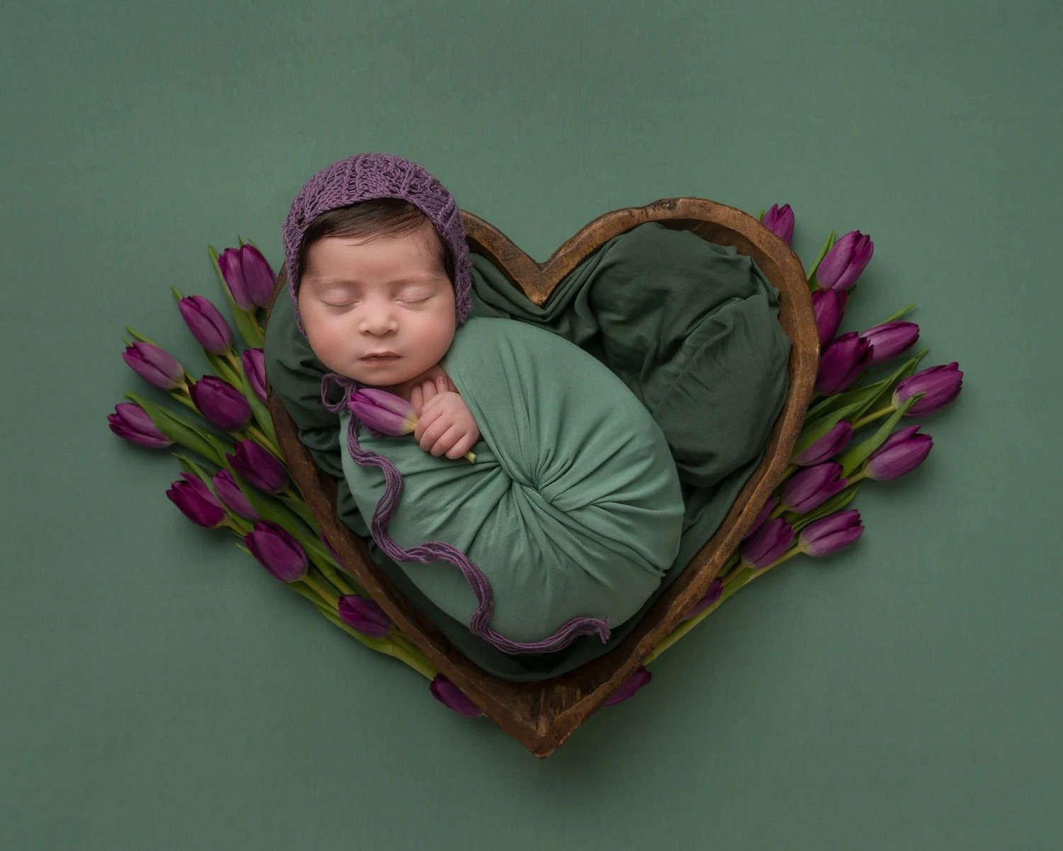 Newborn baby in a purple bonnet asleep in a heart-shaped bowl with tulips, by award-winning newborn photographer Jitka Wenham, Milton Keynes