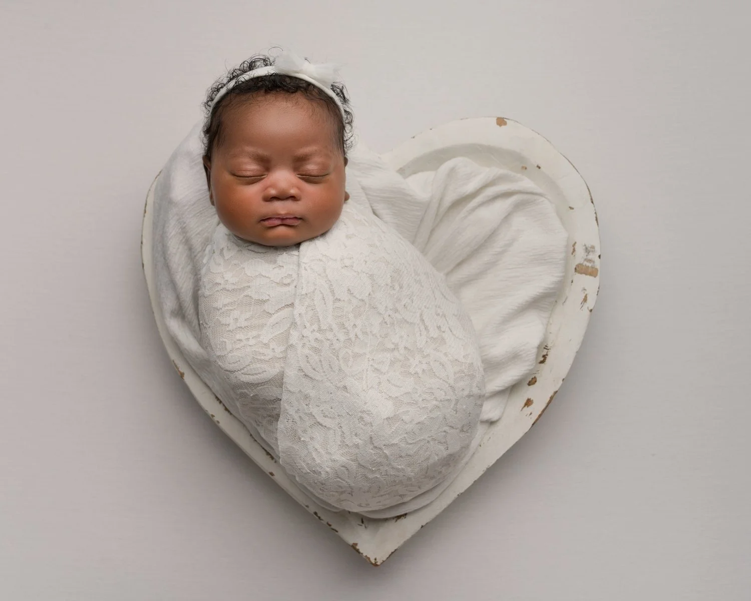 Sleeping newborn baby wrapped in white, posed safely in a white heart bowl at Jitka Wenham Photography, Milton Keynes