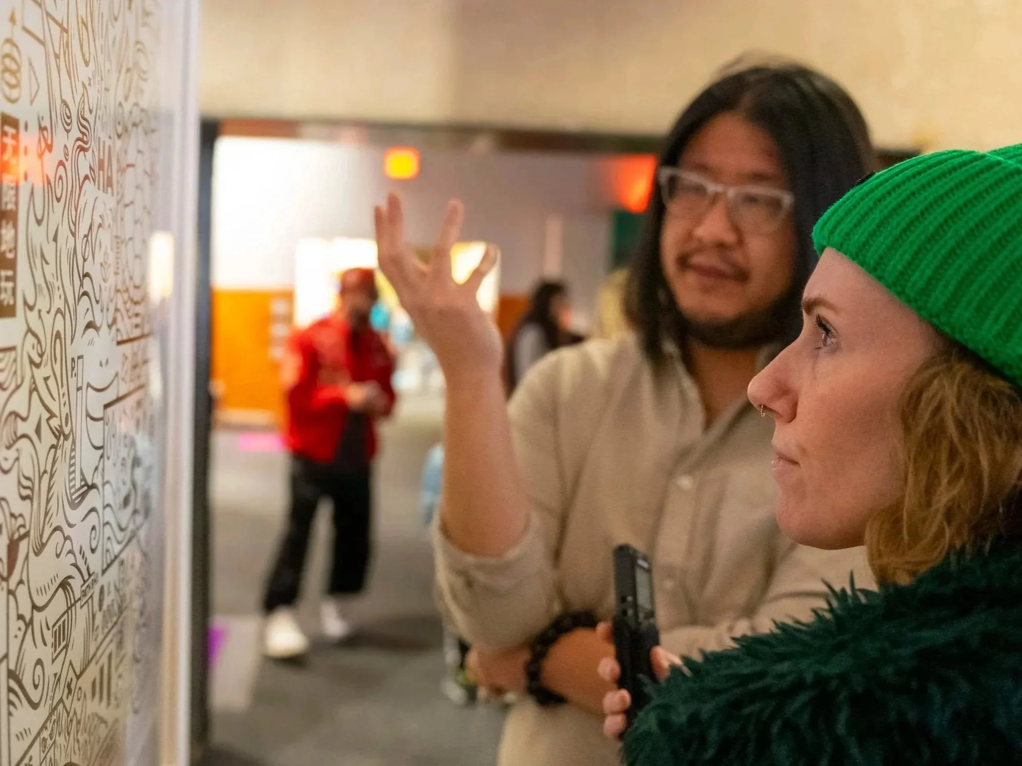 Two people in conversation looking at a wall with artwork, one is gesturing while the other holds a recording device. The woman on the right wears a green beanie and has curly hair, the man on the left wears glasses and a beige shirt.