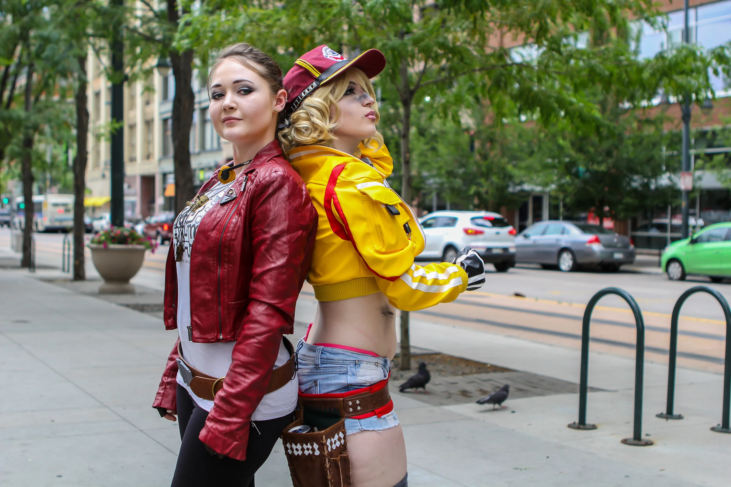 Two young women standing back-to-back on a city sidewalk surrounded by trees and parked cars. One woman wears a red leather jacket and jeans; the other wears a yellow jacket, crop top, and shorts.
