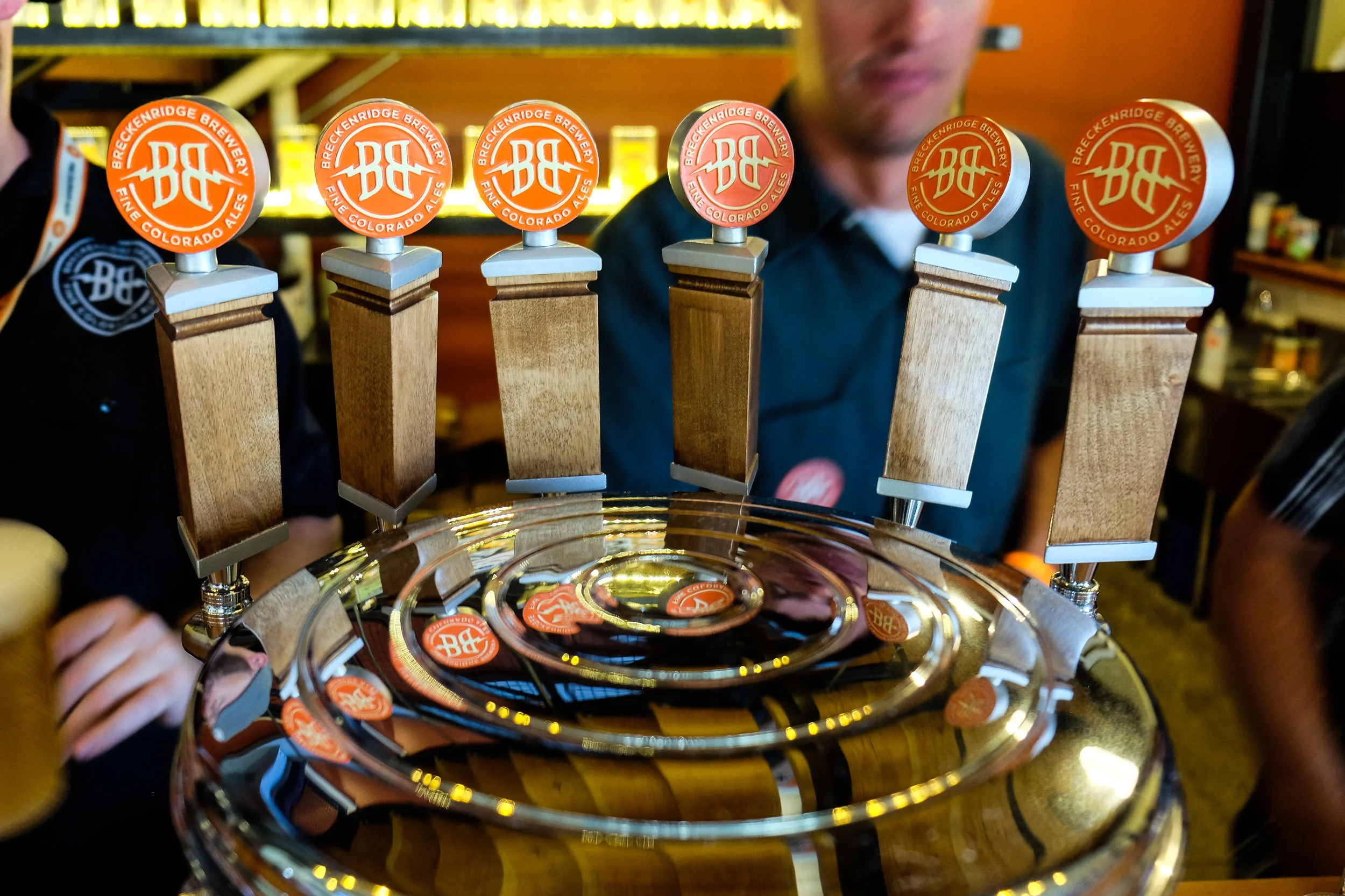 Beer tap handles with Breckenridge Brewery logos lined up on a chrome beer tap. Several people are in the background at a bar or brewery.