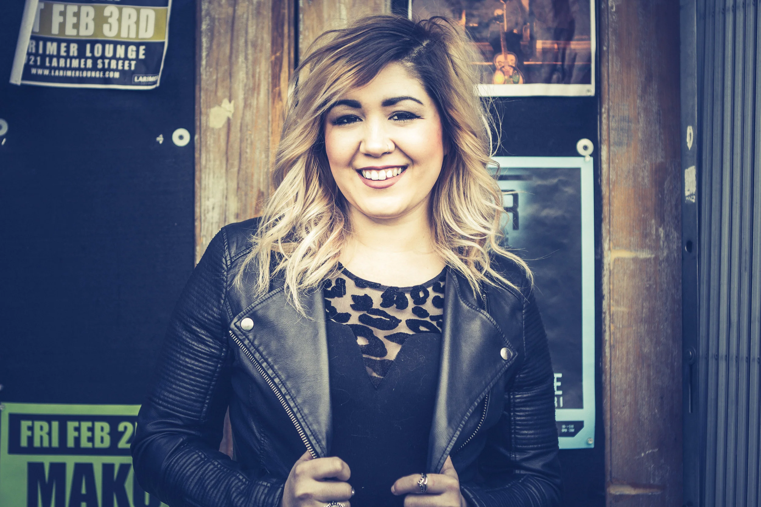 A woman with blonde hair smiling, wearing a black leather jacket and a leopard-print top, standing in front of posters on a wooden wall.