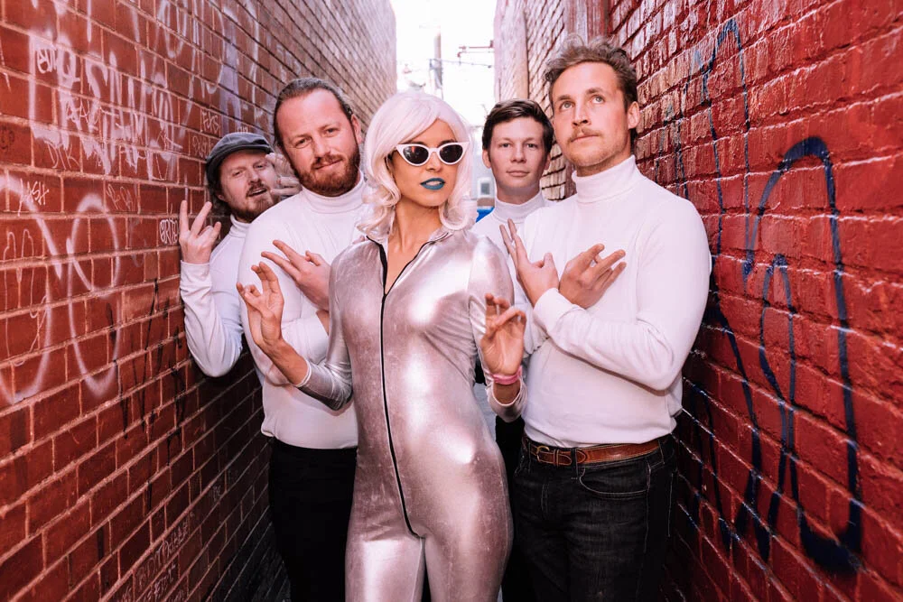 A woman with platinum blonde hair, wearing sunglasses and a shiny silver jumpsuit, posing with four men in a narrow alleyway with red brick walls covered in graffiti.
