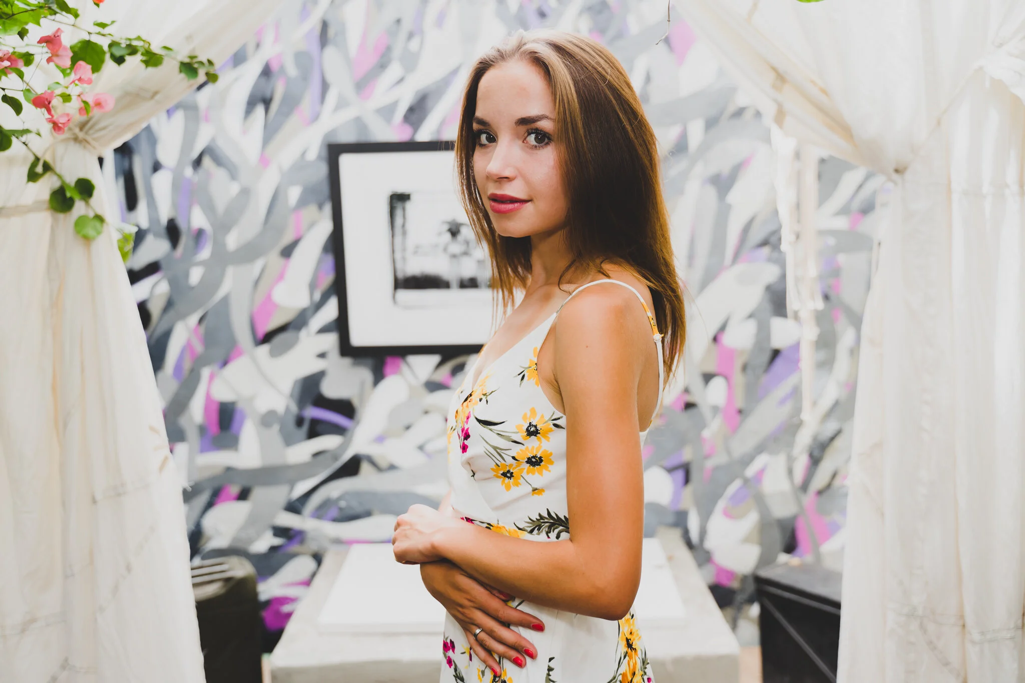 A woman with shoulder-length brown hair in a floral dress posing indoors near a decorative wall and framed picture.