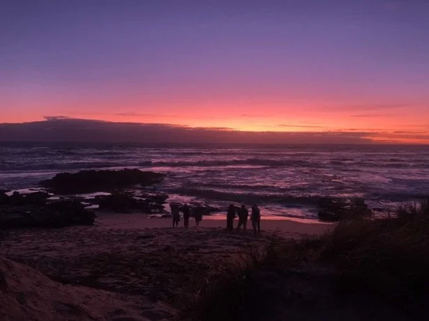A silhouette of six people looking at a purple and orange sunset at a beach in Blairgowrie