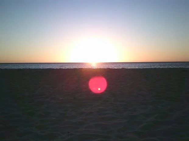 A bright yellow sunset against a pale blue sky at Brighton Beach, Victoria. There is a large pink orb in the middle of the photo