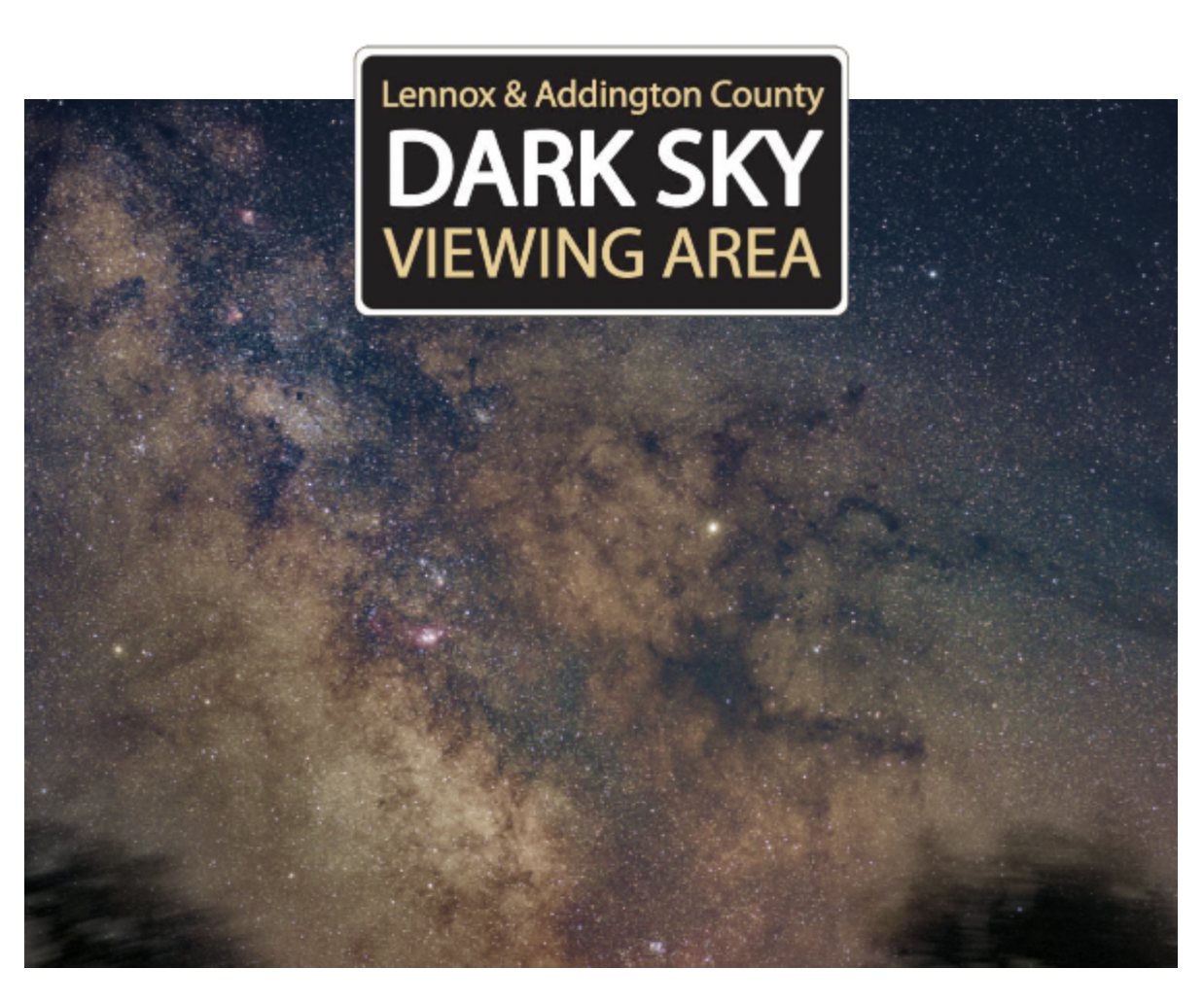 Night sky filled with stars and the Milky Way galaxy over a body of water with tree silhouettes, with a sign reading "Lennox & Addington County Dark Sky Viewing Area."