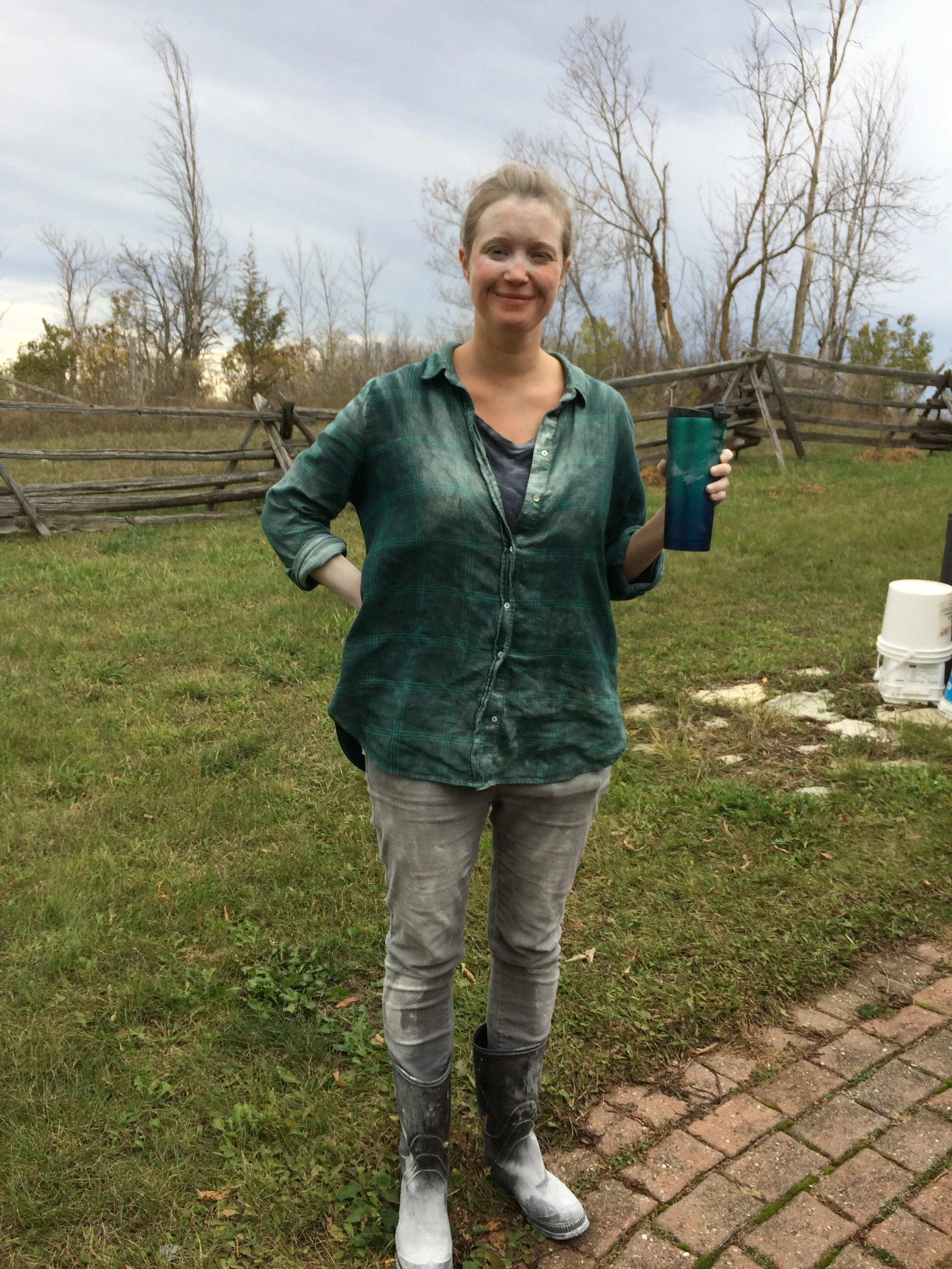 A woman standing outdoors on a grassy field, holding a blue tumbler, wearing a green plaid shirt, gray jeans, and white rubber boots, with a wooden fence and bare trees in the background under cloudy skies.