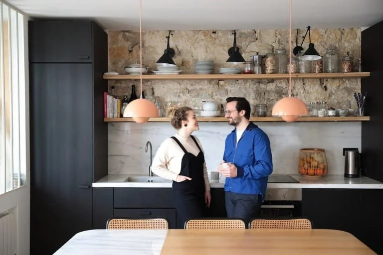 Un homme et une femme discutent dans une cuisine moderne avec une étagère en bois, des suspensions roses, et une gamme noire.