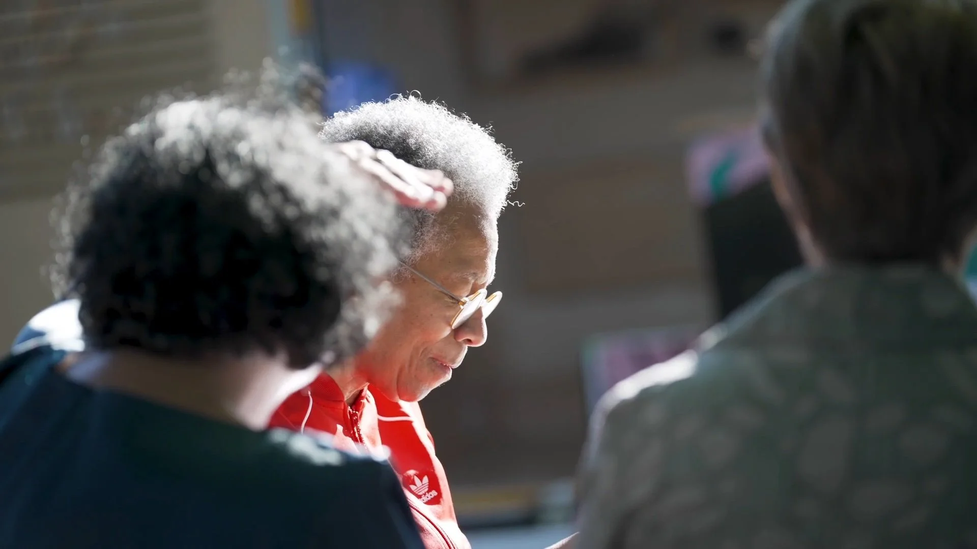 An elderly woman with gray hair and glasses sitting between two people, reading or looking at something, with sunlight illuminating her face.