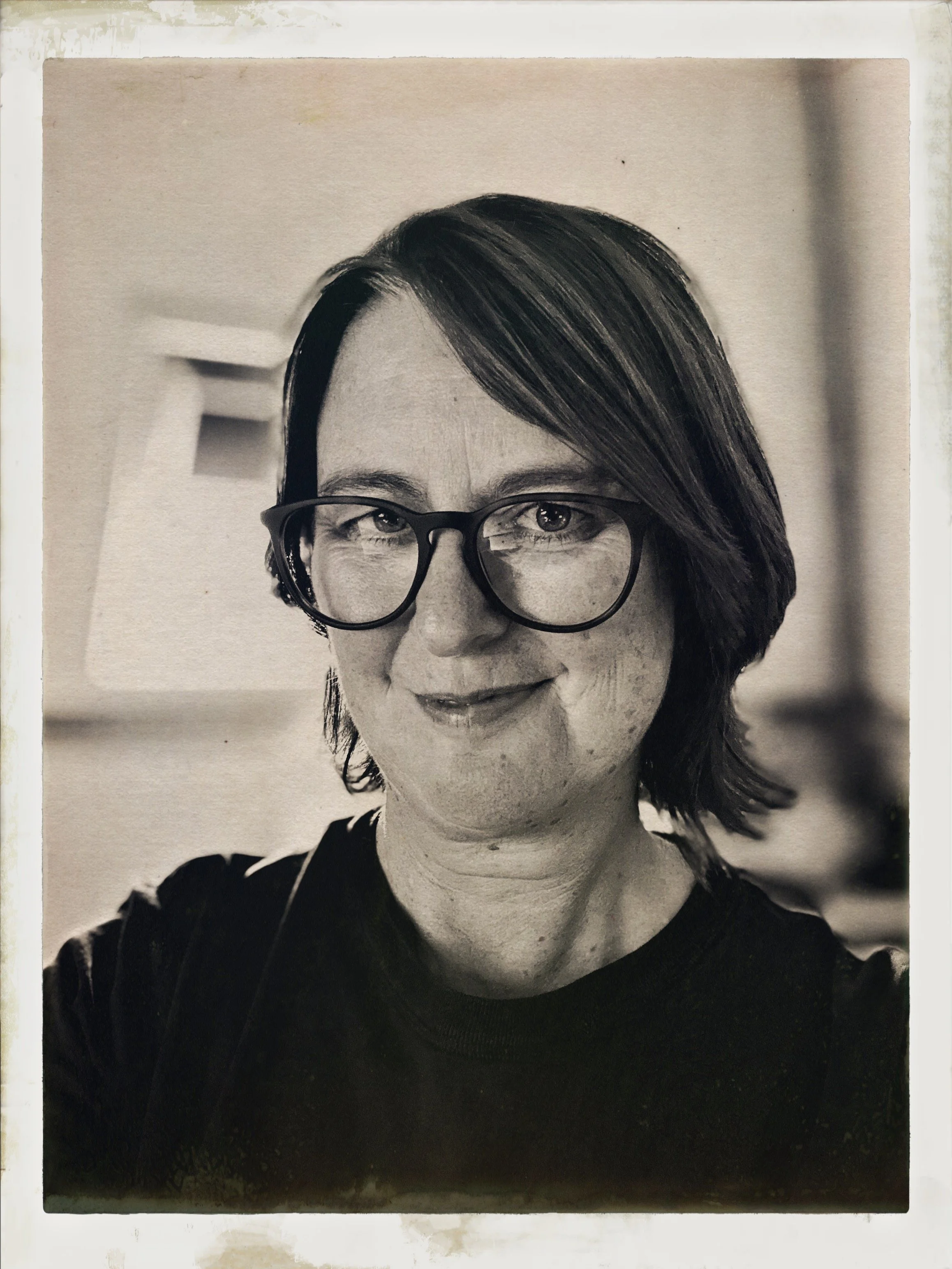 Black and white portrait of a woman with short dark hair, glasses, and a slight smile, indoors with blurred background.