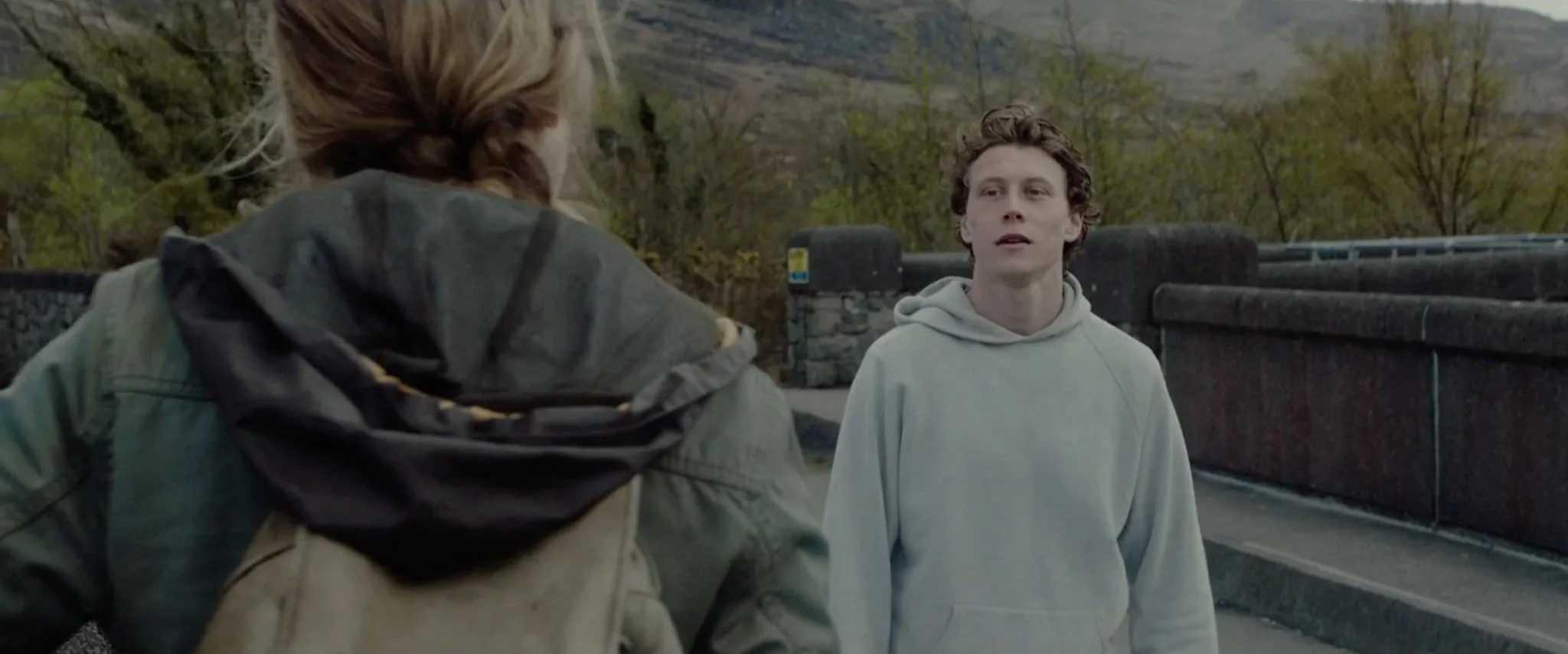 Two young people having a conversation outdoors on a bridge, with trees and hills in the background.