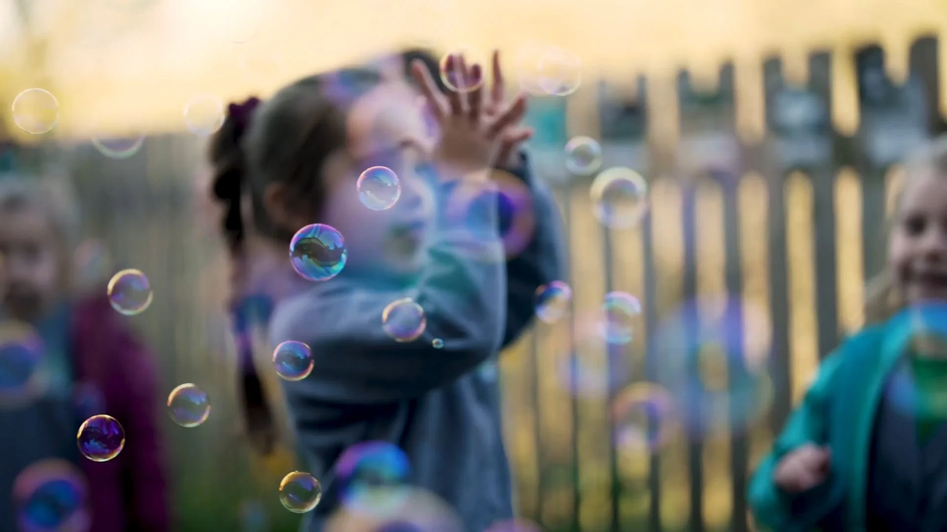 Young children playing outdoors near a wooden fence with bubbles floating in the air, some children raising their hands.
