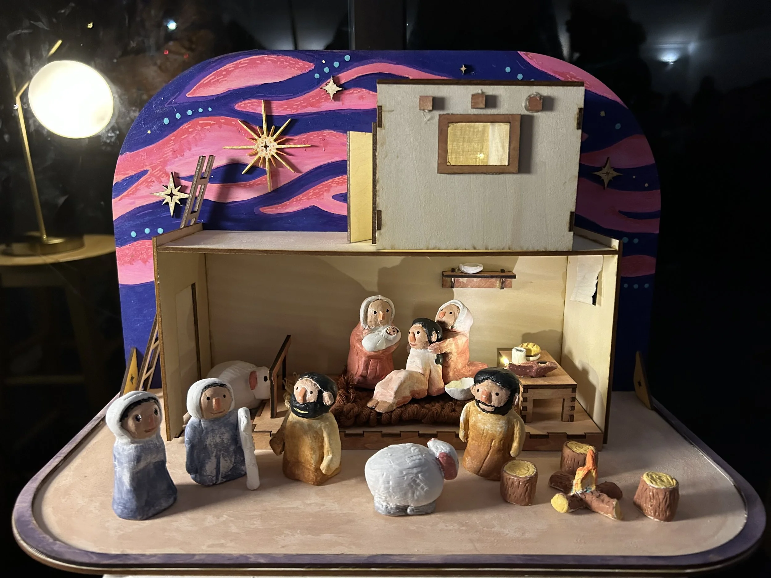 Two-story wooden nativity diorama with a hand-painted night sky mural in pink and purple, sculpted and painted clay figures of Mary holding the newborn Jesus surrounded by attending women, shepherds, and animals gathered around a manger.